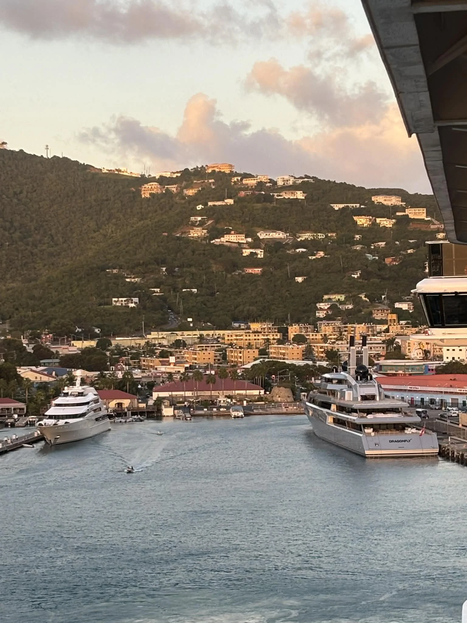 A harbor scene at sunset with boats docked, including a large yacht named Dragonfly, and a hillside with houses and trees in the background.