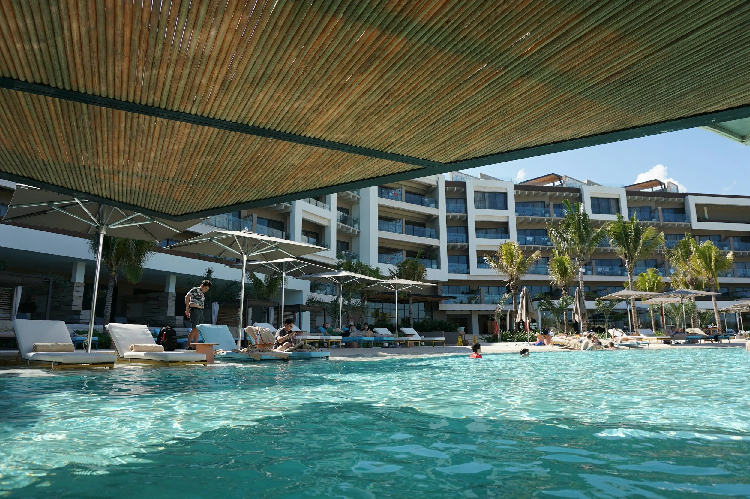 Swimming pool area at a hotel with lounge chairs, umbrellas, and palm trees, with a multi-story building in the background on a sunny day.