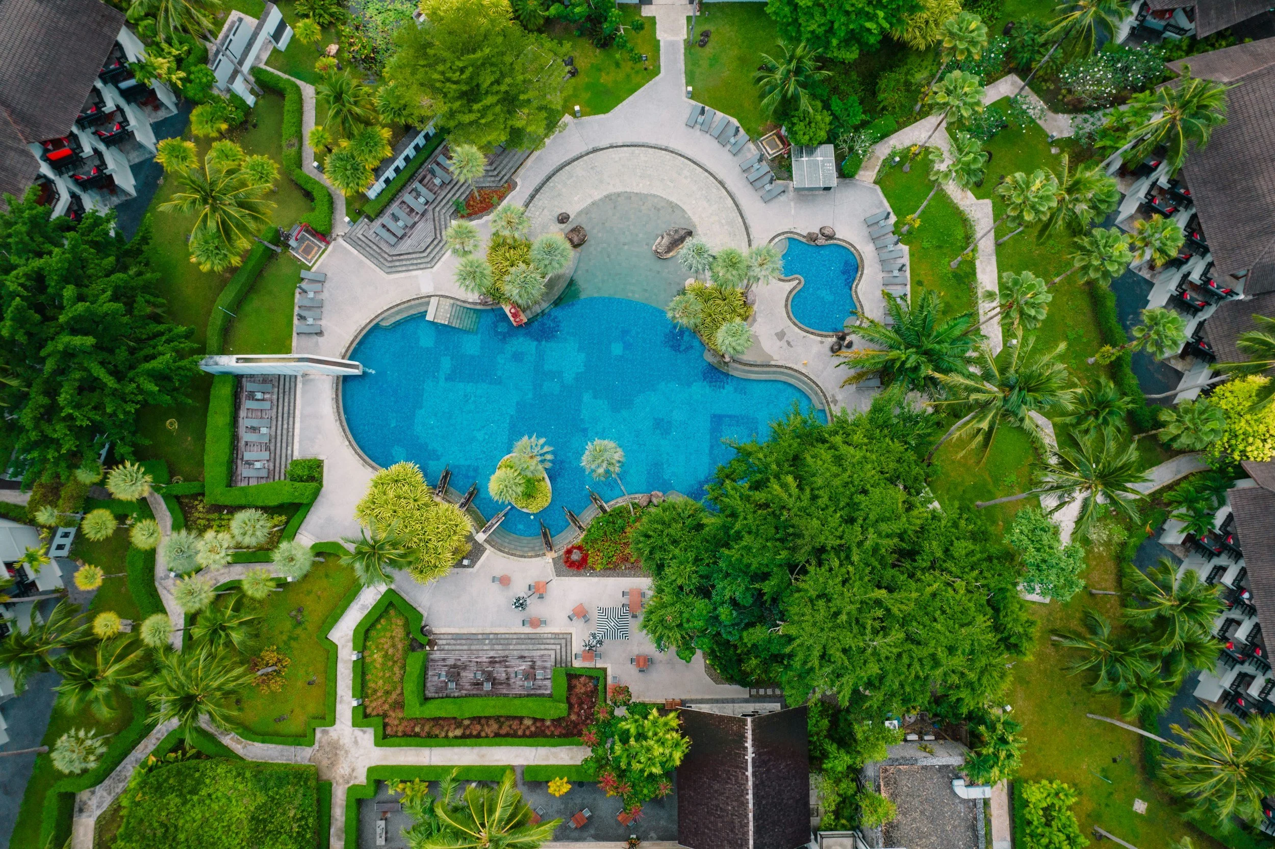 Aerial view of a swimming pool area at a tropical resort surrounded by lush green trees, palm trees, and landscaped gardens. The pool has a curved shape with a small adjacent hot tub and a waterslide. Lounge chairs and pathways are visible around the pool, with resort buildings and multi-story villas bordering the area.