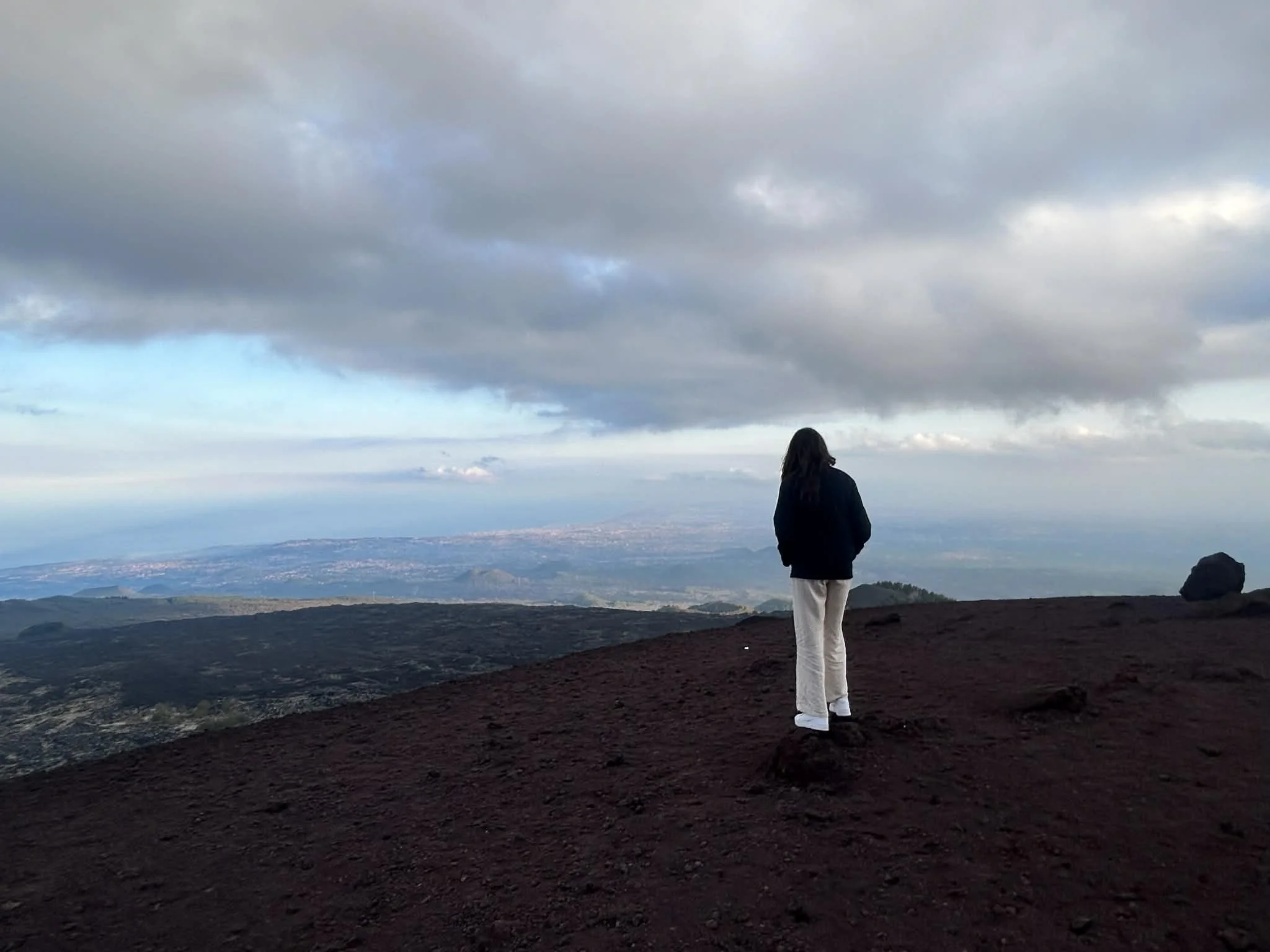 A woman standing on a dark rocky hilltop, overlooking a vast landscape with cloudy skies and distant mountains or hills.