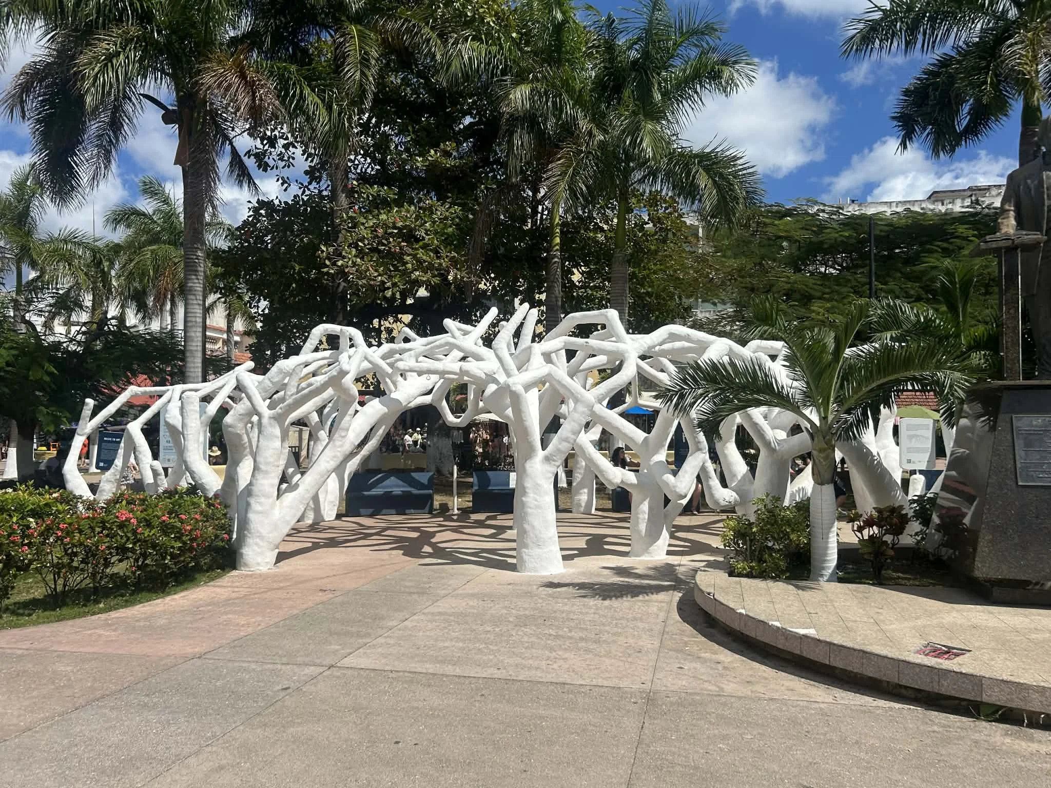 Outdoor art installation of white painted tree sculptures with intertwined branches, surrounded by palm trees and greenery in a park setting on a sunny day.