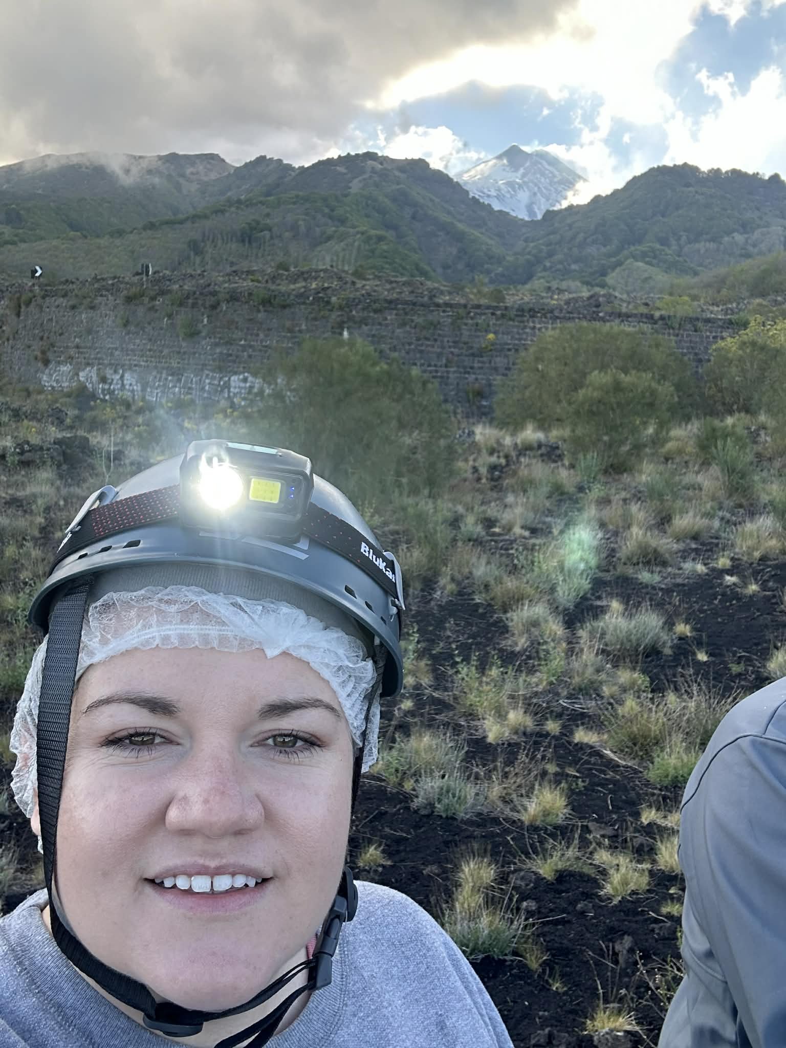 A woman wearing a helmet with a headlamp takes a selfie outdoors in a mountainous area with lush green hills, volcanic terrain, and snow-capped peaks in the background.