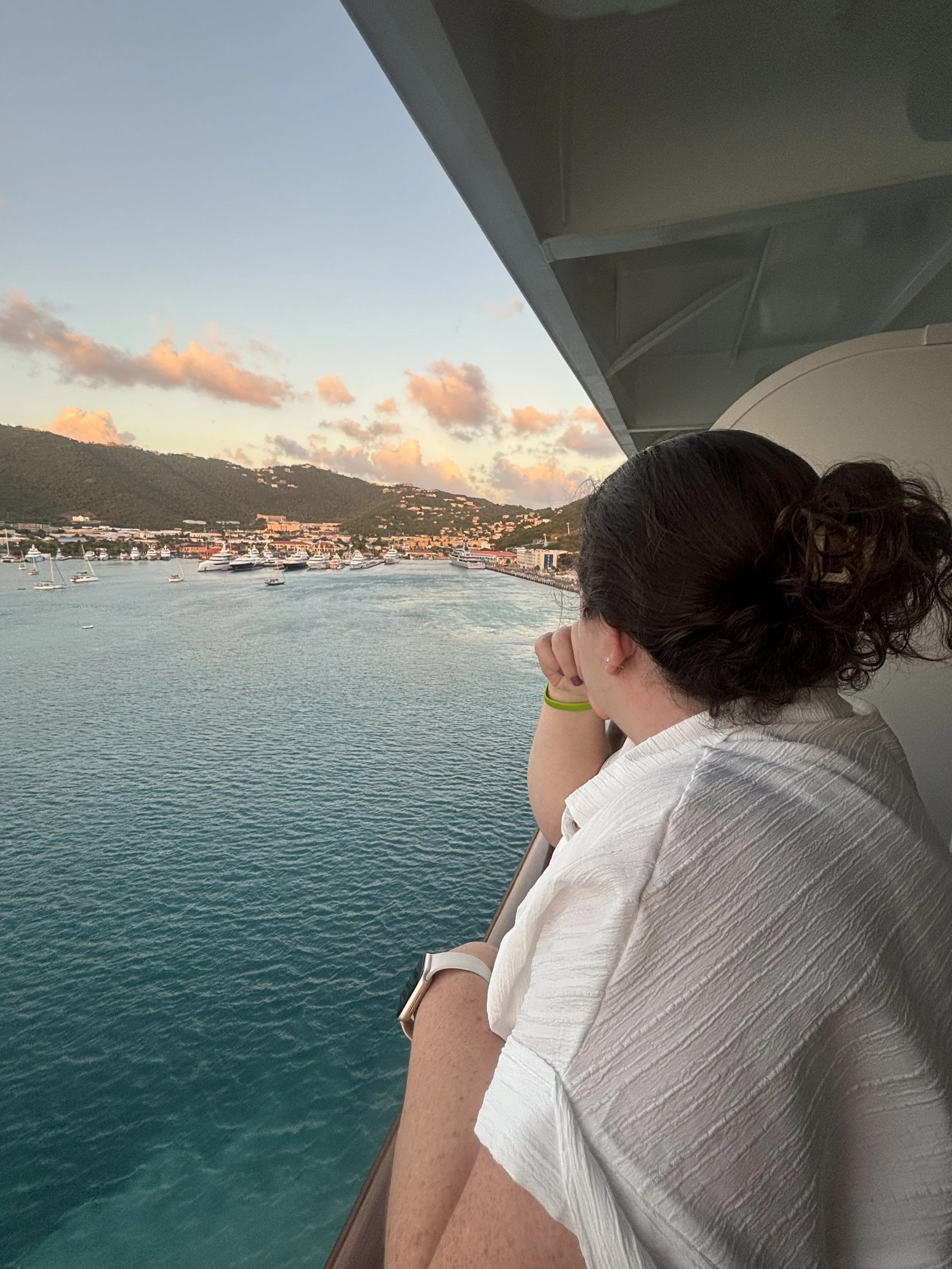 A woman with dark hair tied back, wearing a white shirt and a yellow wristband, looks out of a window at a harbor with boats and a scenic backdrop of hills with buildings at sunset.