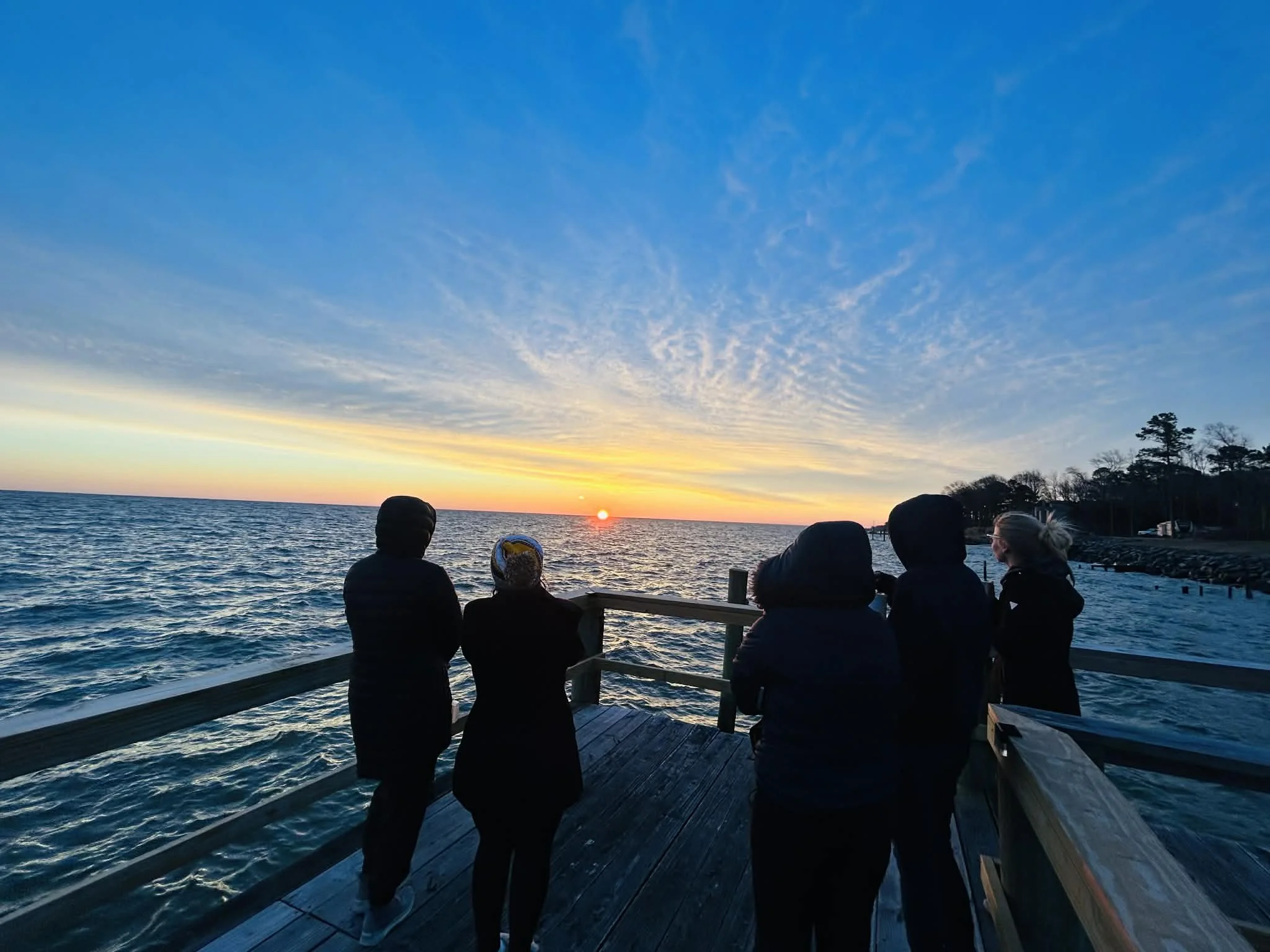 Five people standing on a wooden pier watching a sunset over the ocean.