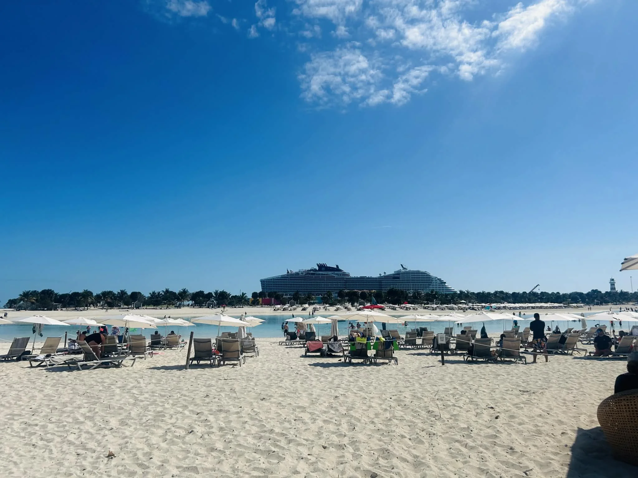 Beach with white umbrellas and lounge chairs, people relaxing and walking, large cruise ships in the background, clear blue sky with a few clouds.