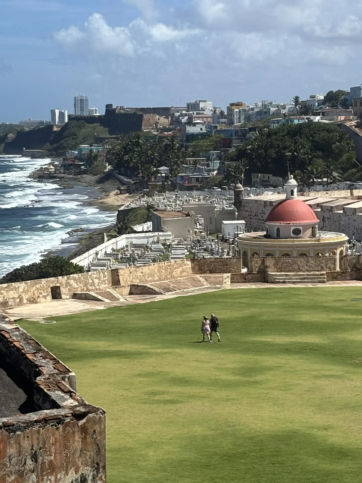 Two people walking on a grassy field with historic buildings, a red-domed chapel, and a coastline with waves in the background.