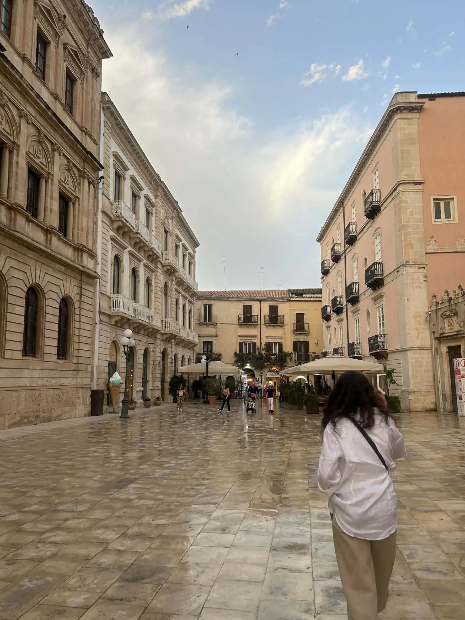 A woman walking on a wet stone-paved street in a European city with historic buildings, cafes, and umbrellas, under a partly cloudy sky.