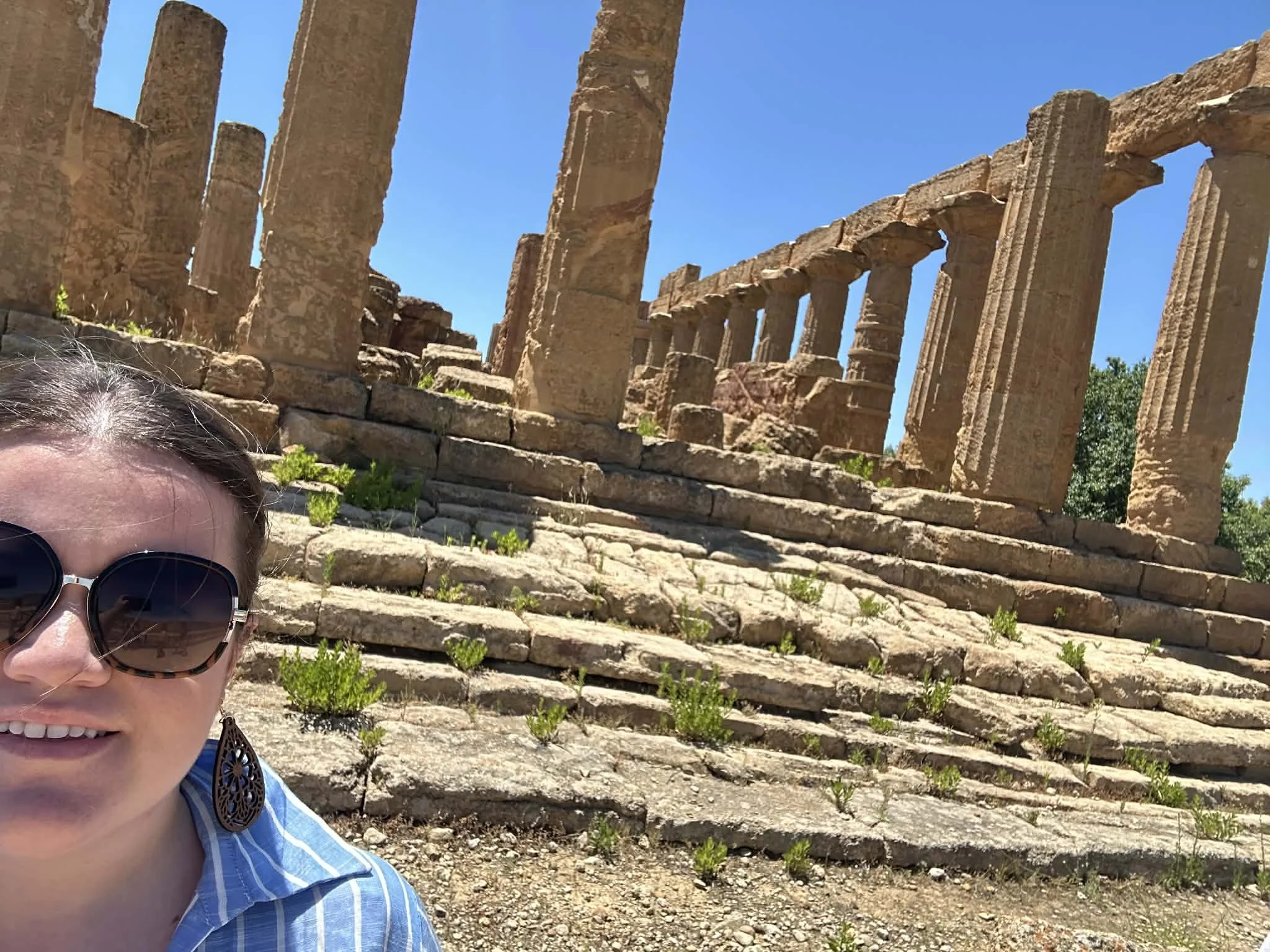 A woman smiling wearing sunglasses and earrings, standing in front of ancient stone ruins with columns and steps, under a clear blue sky.