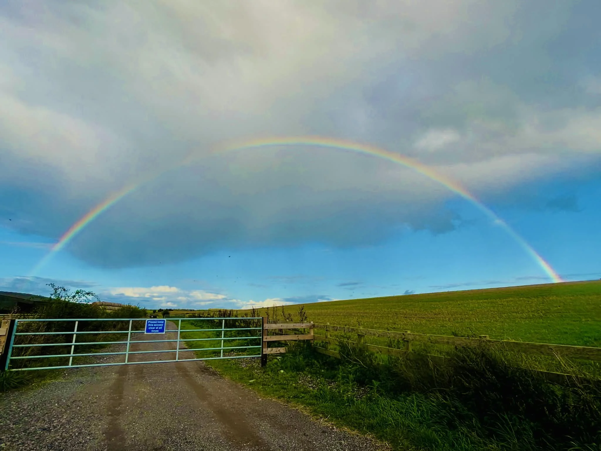 A rainbow arching across a cloudy sky over a grassy field, with a gravel farm or country road in the foreground and a blue gate with a sign that says 'Please keep gates closed'.