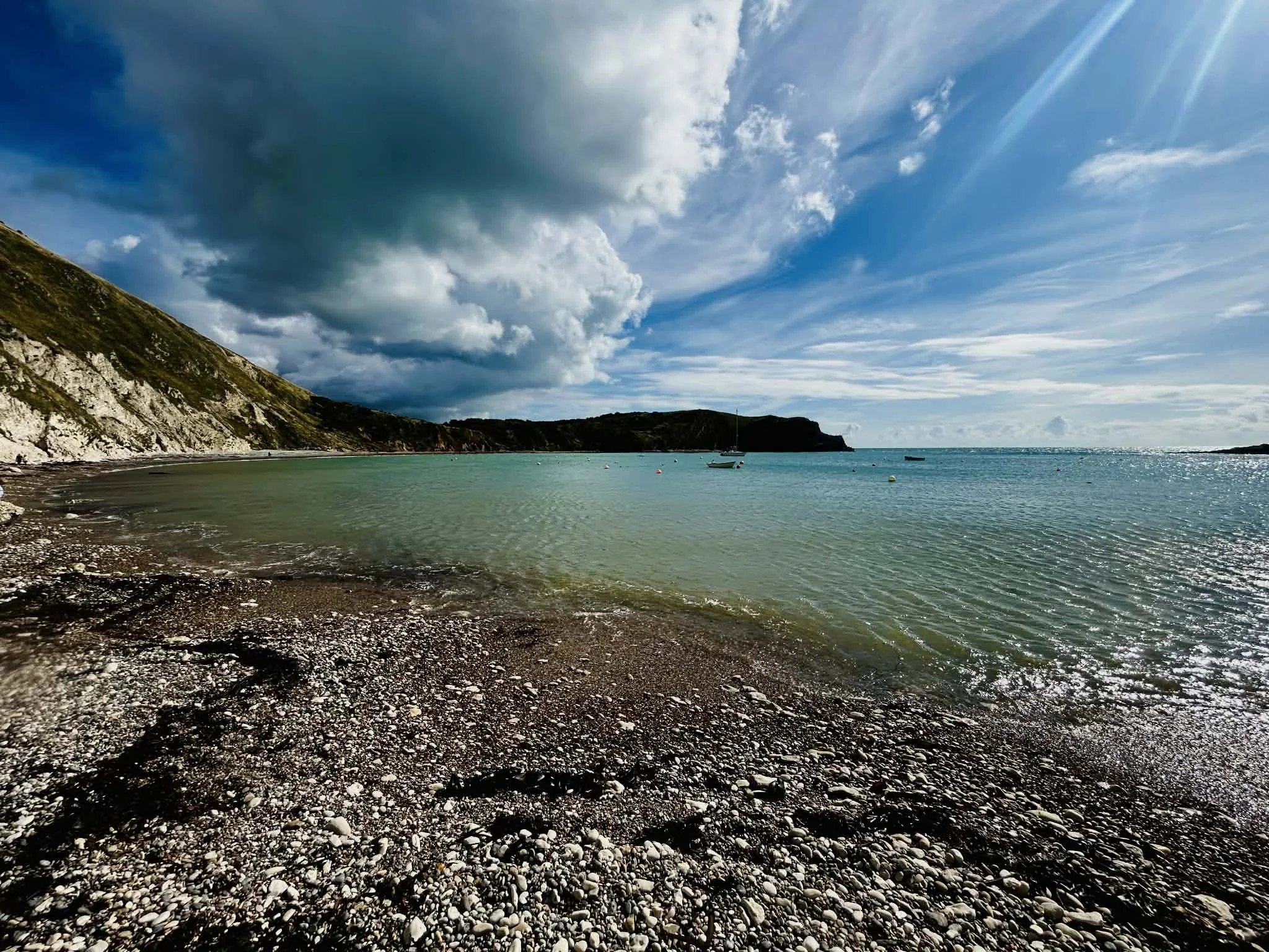 Coastal scene featuring a pebble beach, calm green waters, a sailboat, and a partly cloudy sky with sunlight rays.