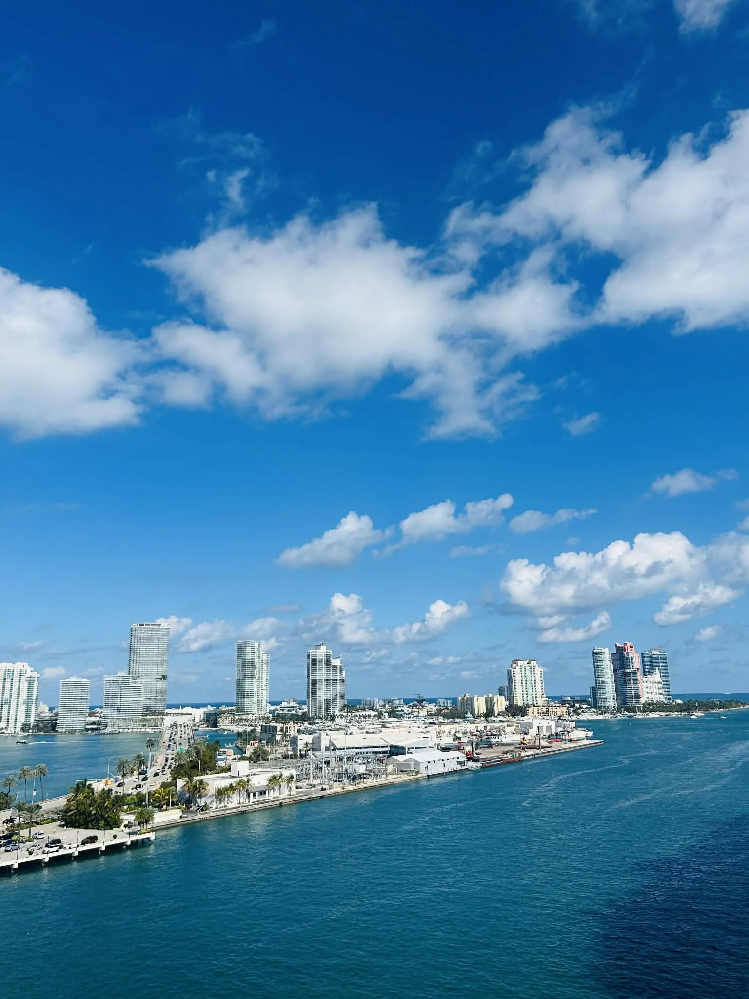 City skyline with high-rise buildings along a waterfront under a partly cloudy blue sky.