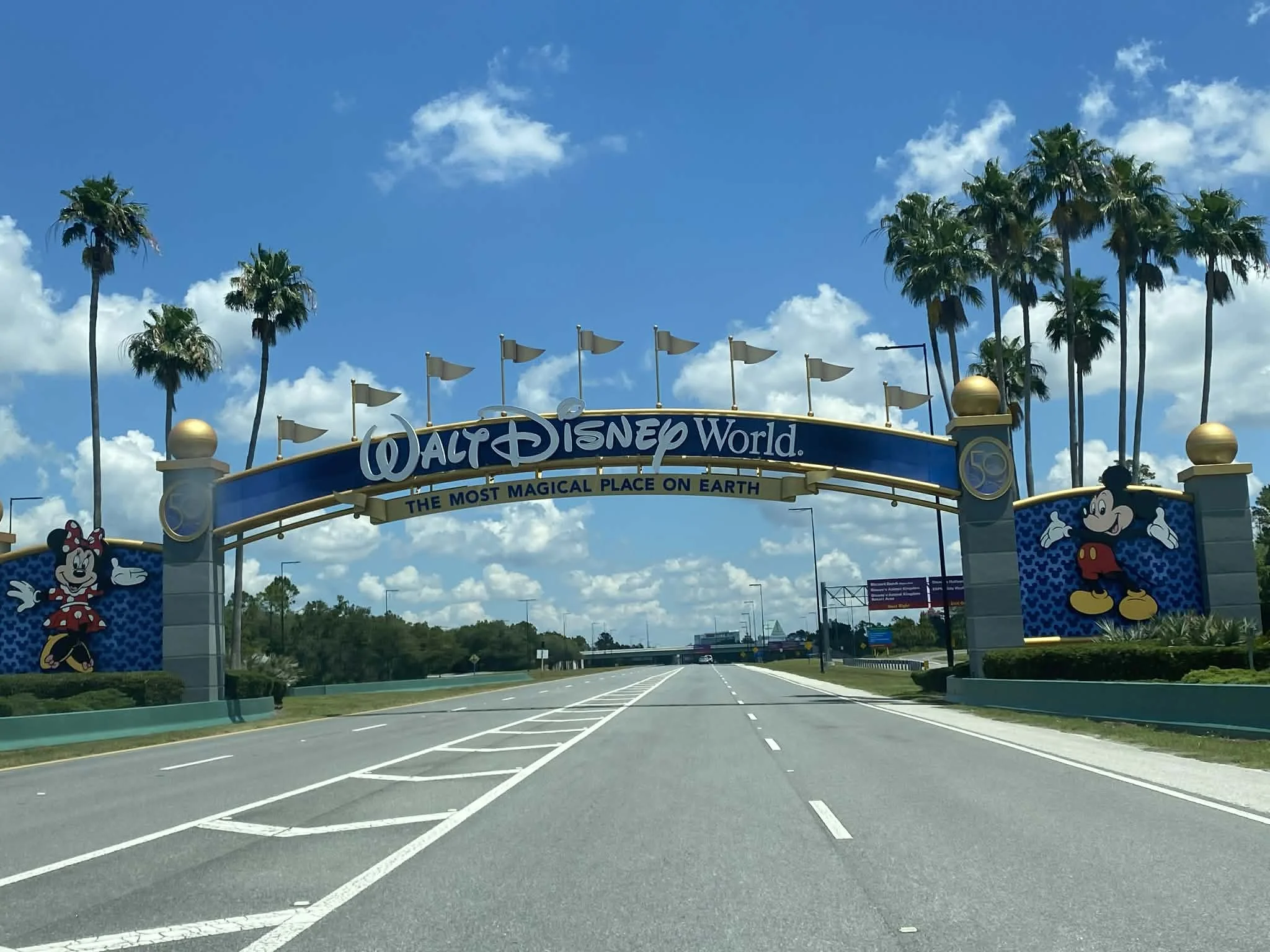 Entrance sign to Walt Disney World with Mickey Mouse and Minnie Mouse illustrations, palm trees, blue sky, and white clouds.