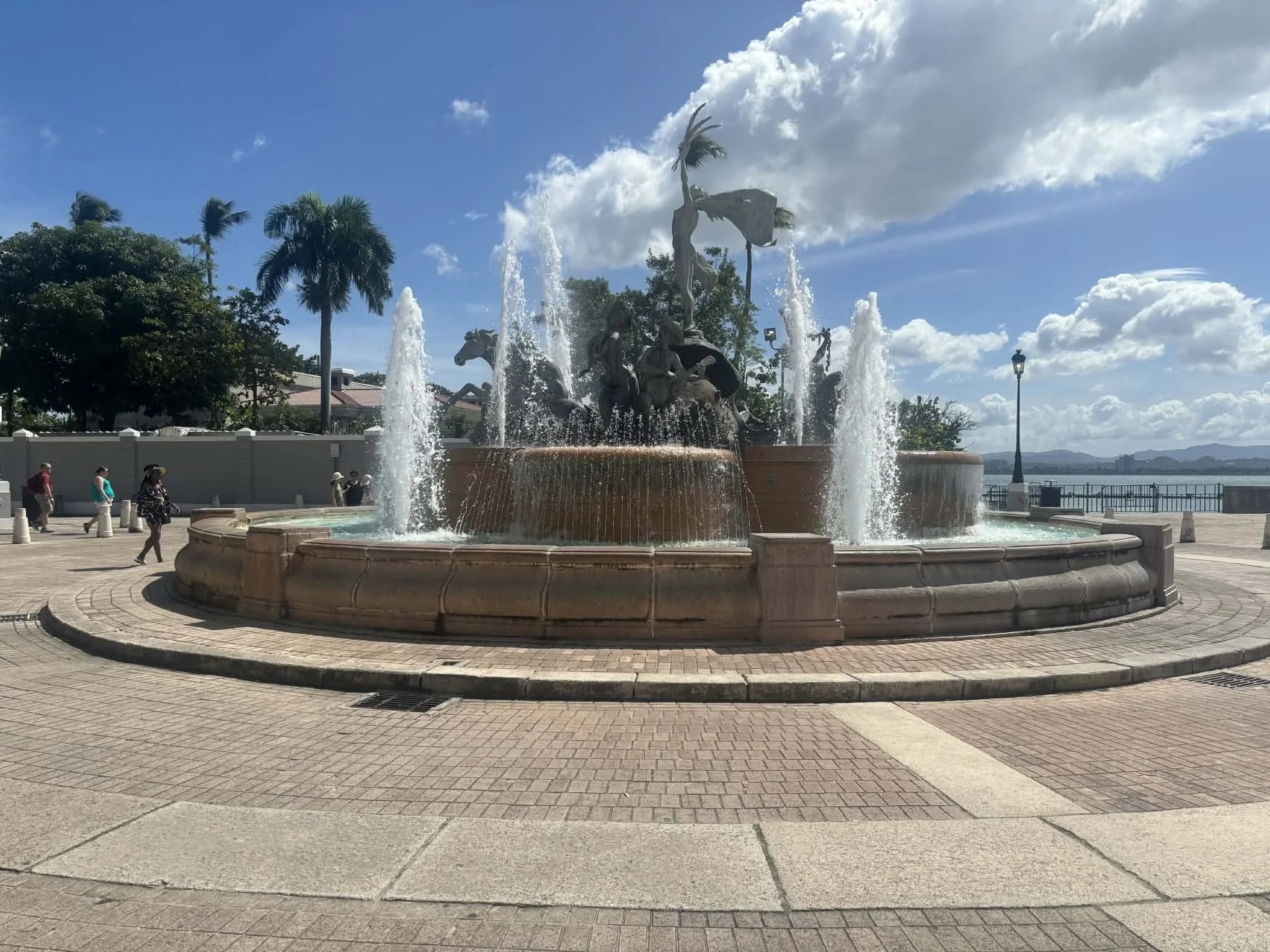A decorative fountain featuring sculptures of horses and a person with water jets, located on a waterfront promenade with people walking and trees in the background.