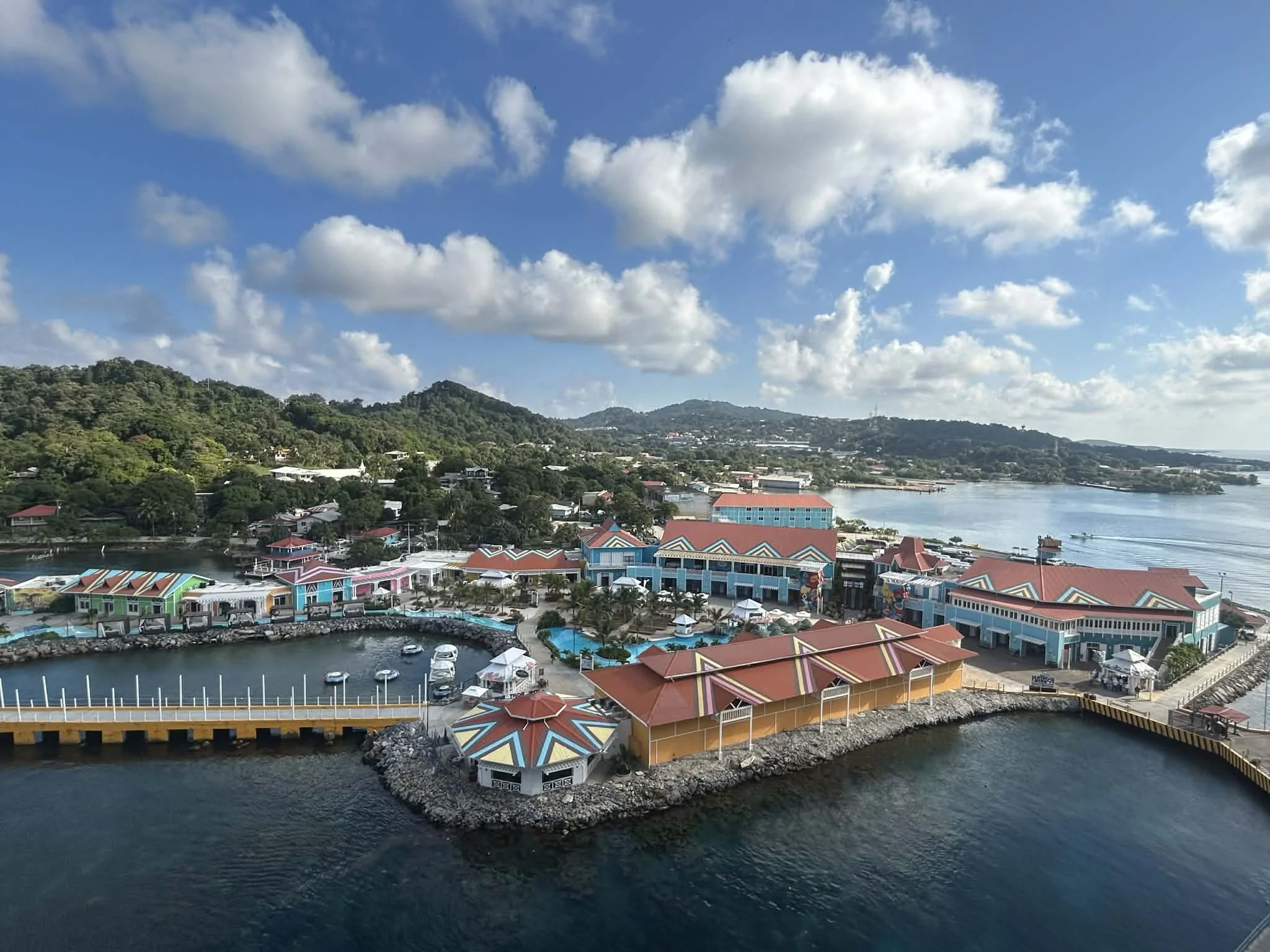 Aerial view of a seaside town with colorful buildings, a marina with boats, a body of water, green hills in the background, and a partly cloudy sky.
