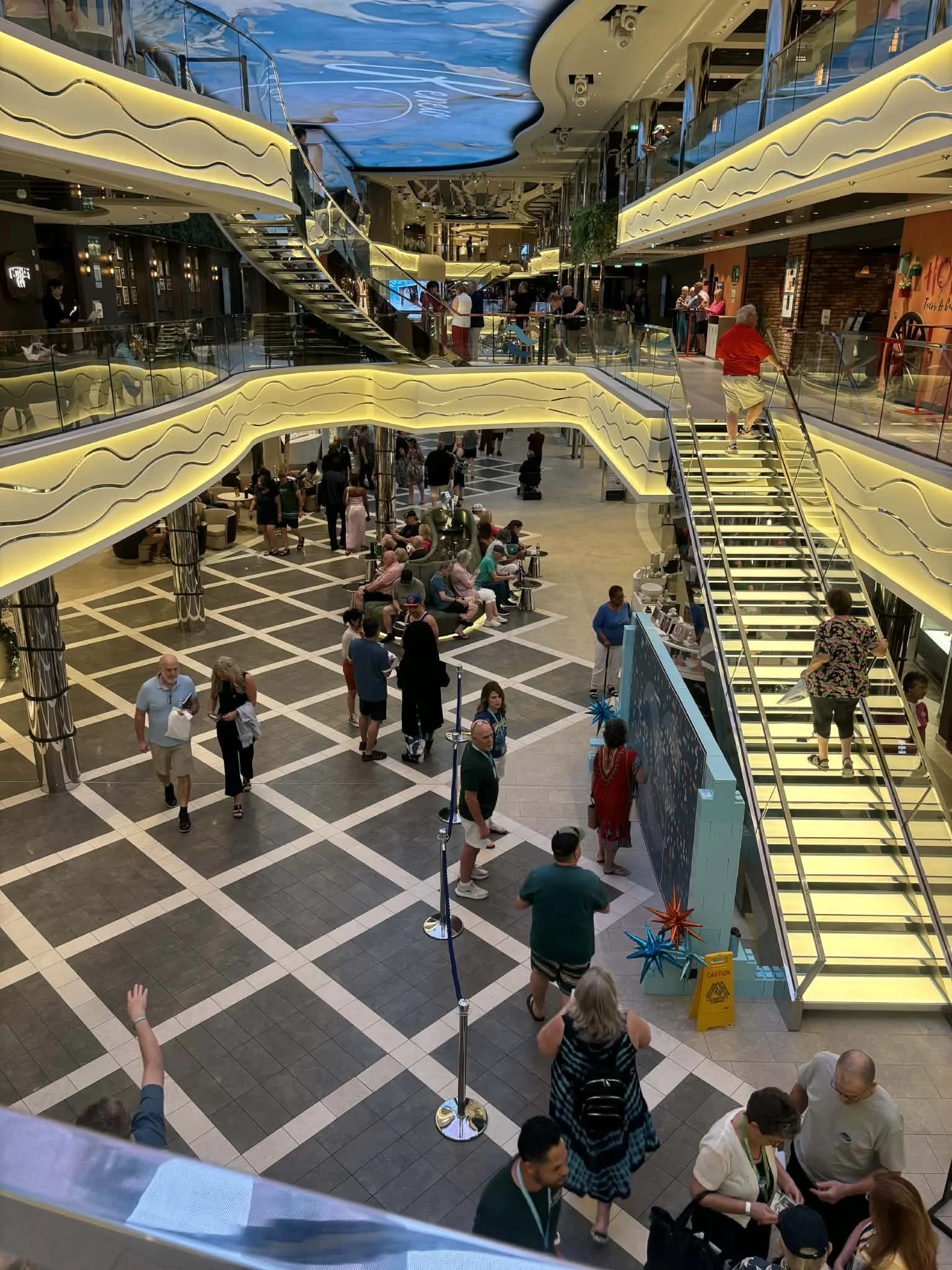 Interior of a shopping mall or cruise ship with multiple floors, escalators, people walking, sitting, and lining up, decorative lighting, and a ceiling mural of a sky with clouds.