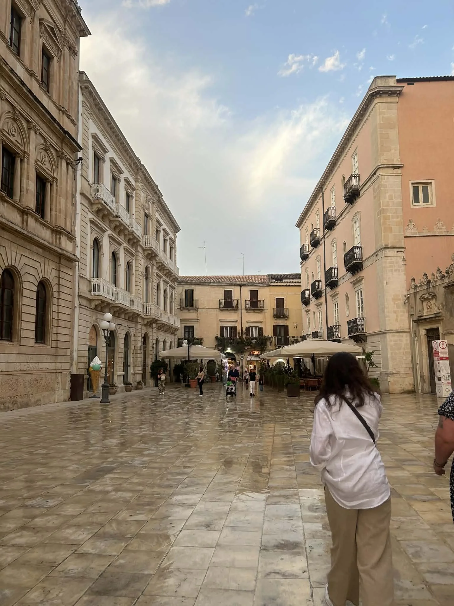 A woman with dark hair, wearing a white blouse and tan pants, stands on a wet cobblestone plaza with her back to the camera. The plaza is surrounded by historic buildings with ornate architecture and small balconies. There are outdoor cafe tables wit