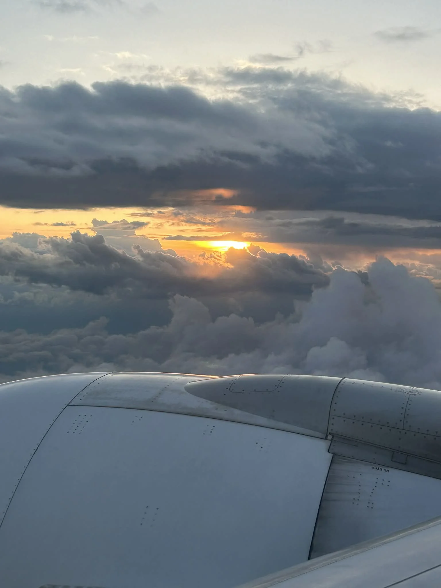 A view from an airplane window showing the airplane engine in the foreground and a cloudy sky at sunset in the background.