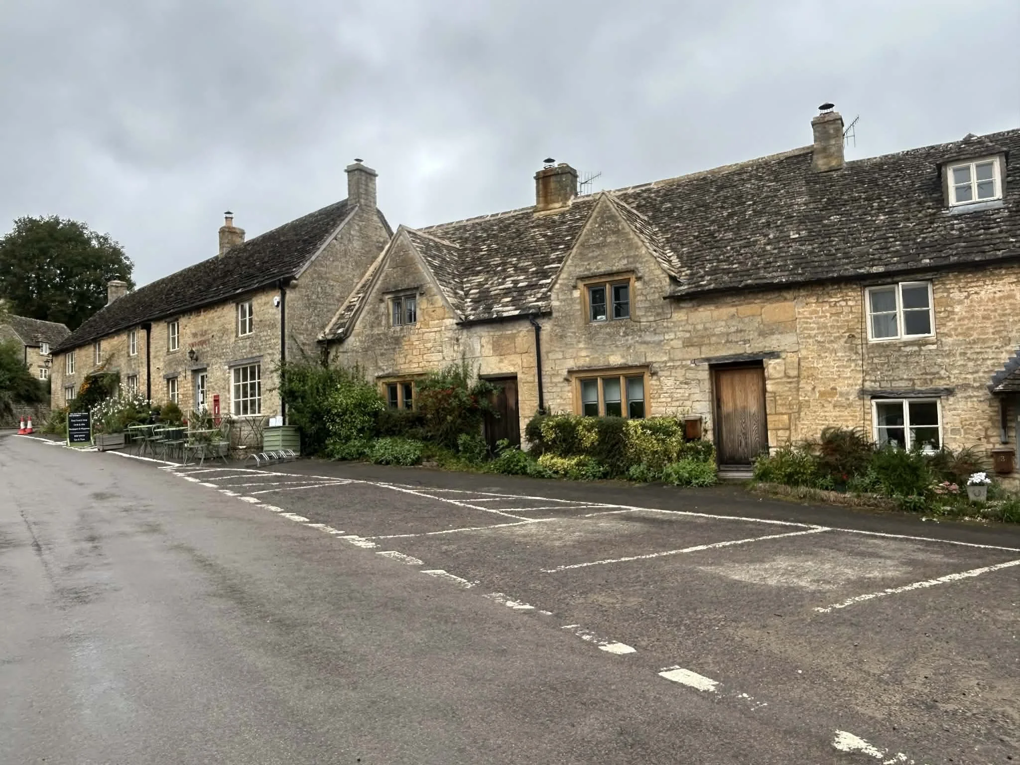 A row of historic stone cottages with pitched roofs and chimneys along a wet road with some parking spaces. The sky is overcast.