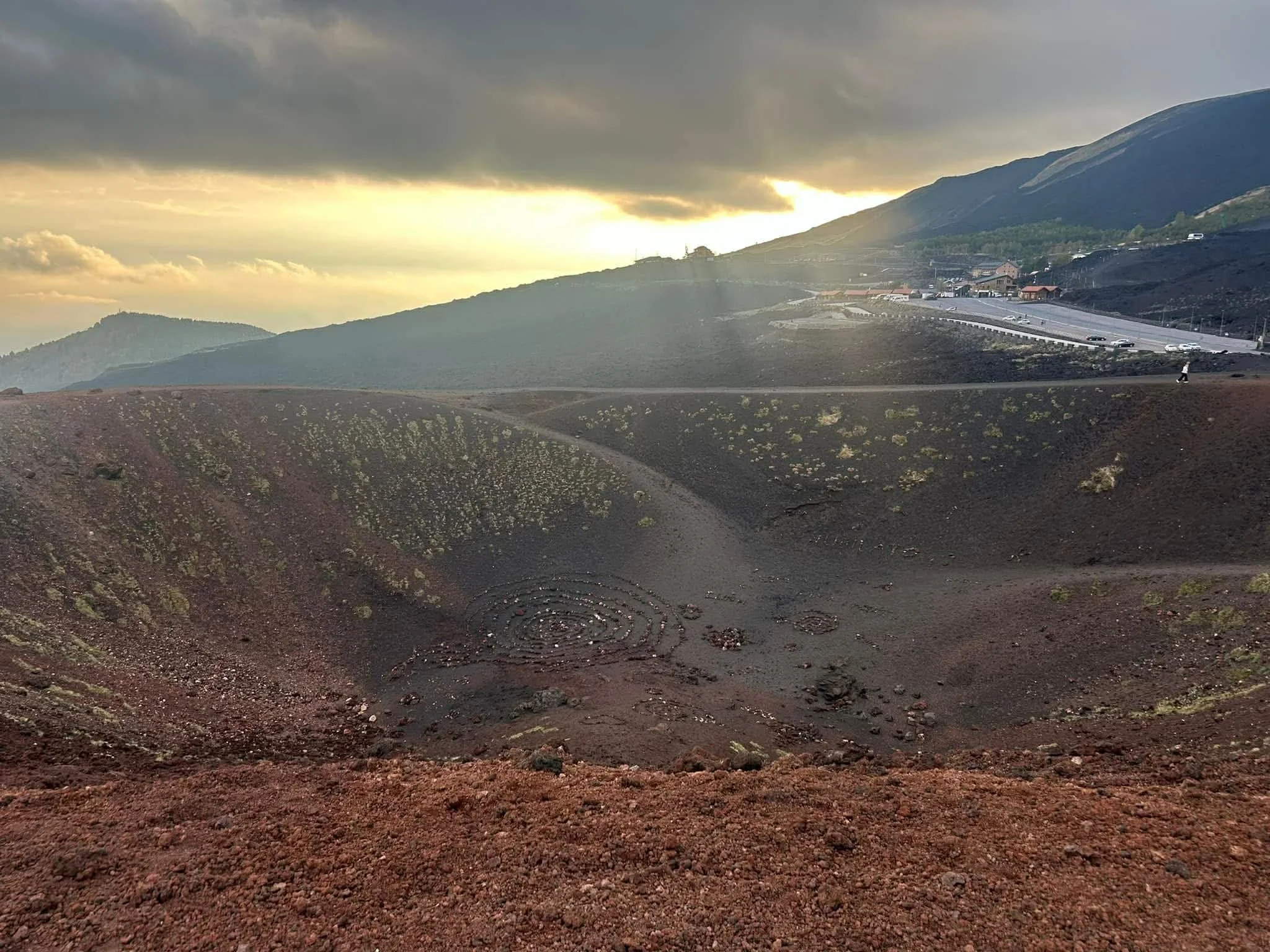 Volcanic crater with reddish-brown soil and sparse vegetation, surrounded by hilly terrain, with a dark cloudy sky and a distant road with cars.