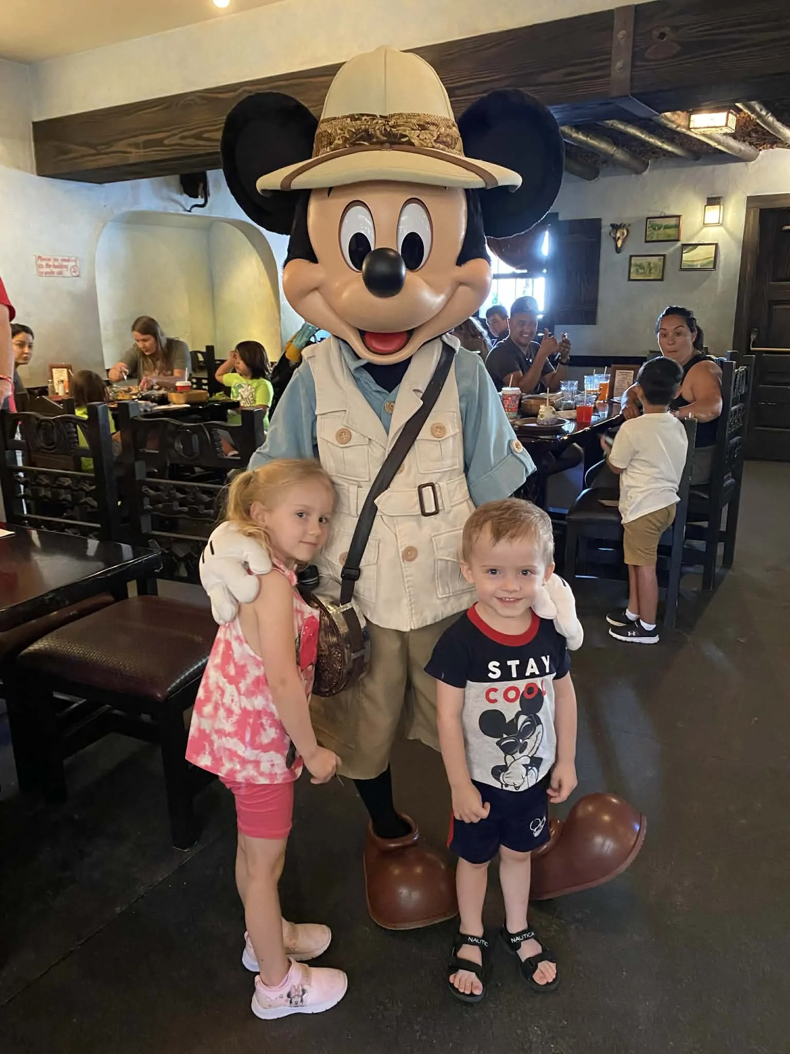 Children posing with Mickey Mouse dressed as an explorer in a restaurant, with other diners in the background.