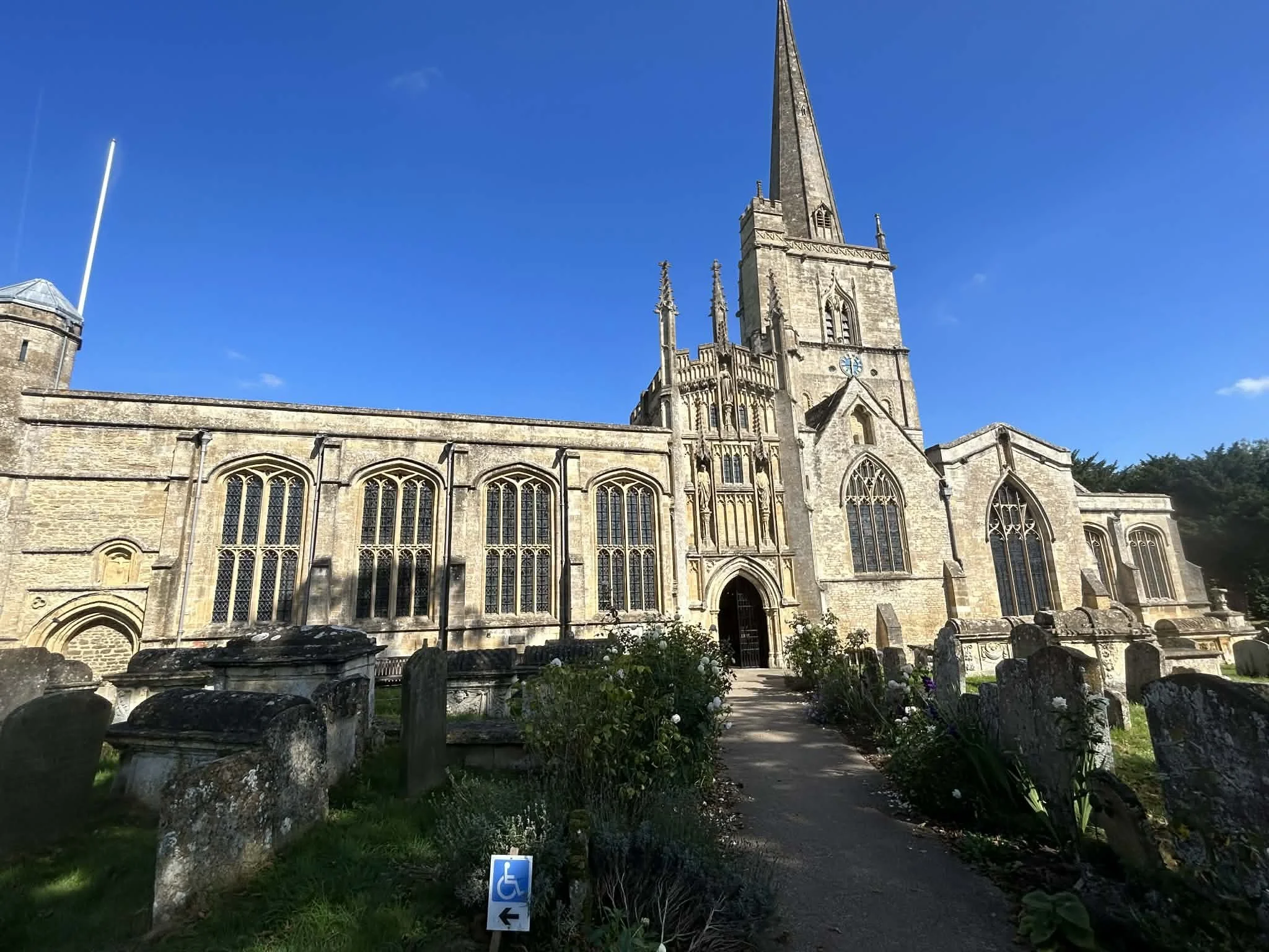 A large stone church with tall windows and a prominent steeple, surrounded by a graveyard with old, weathered headstones, under a clear blue sky.