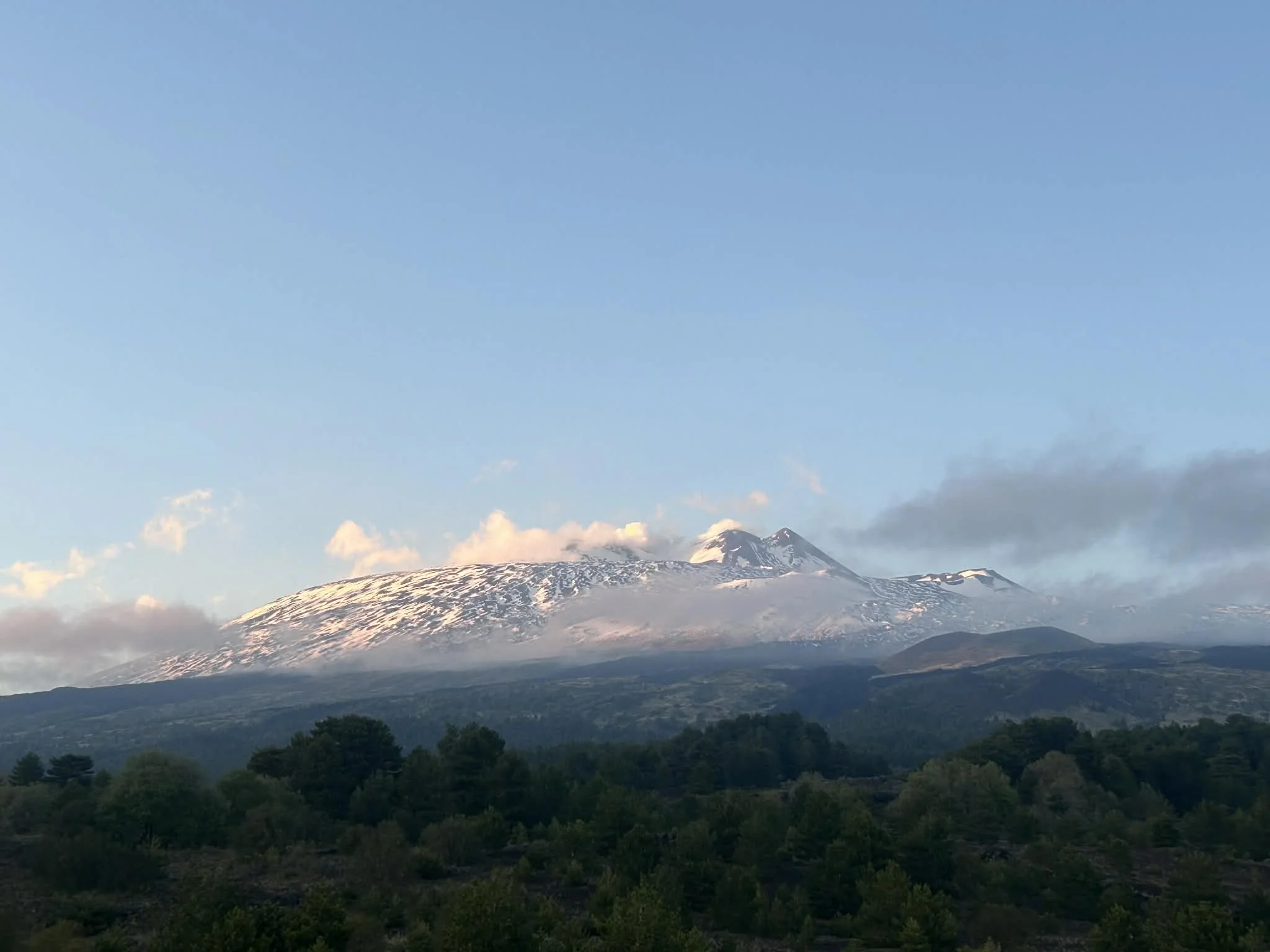 Snow-capped mountain with green forest in foreground, partly cloudy sky with blue background.