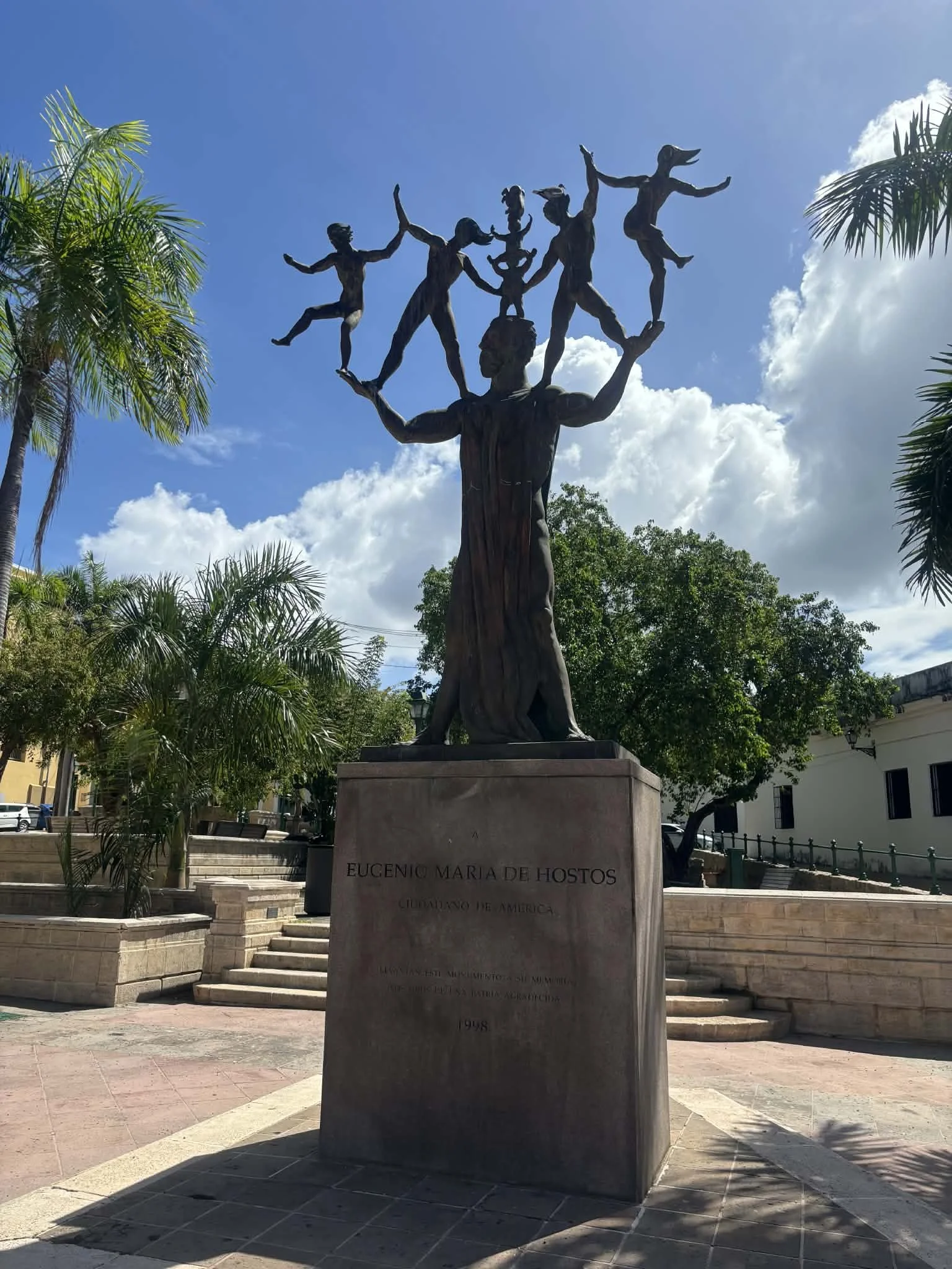 Statue of Euclides Maria de Hostos with children on her arms in a park, surrounded by palm trees and green foliage, under a partly cloudy sky.
