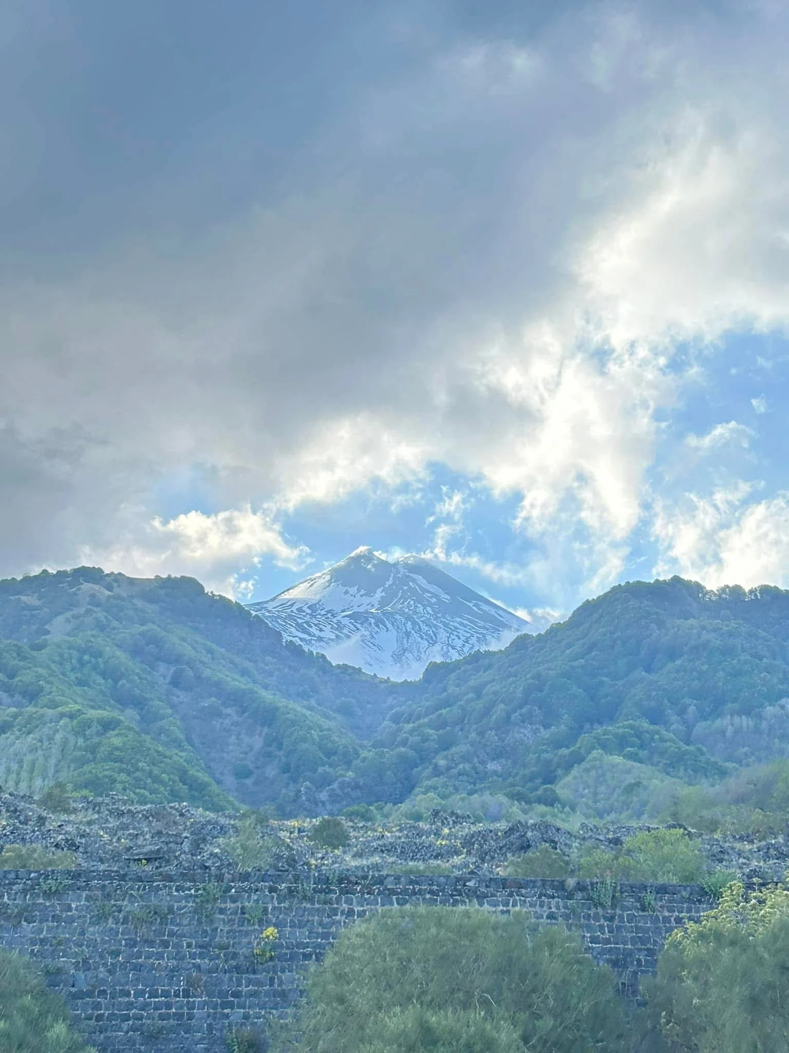 Snow-capped mountain peak surrounded by green hills and cloudy sky.