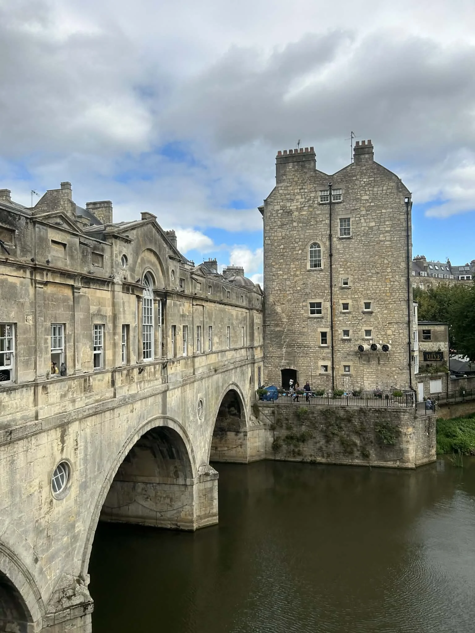 Historic stone buildings over a river in a city, with a bridge and people walking on a walkway by the water, under a cloudy sky.