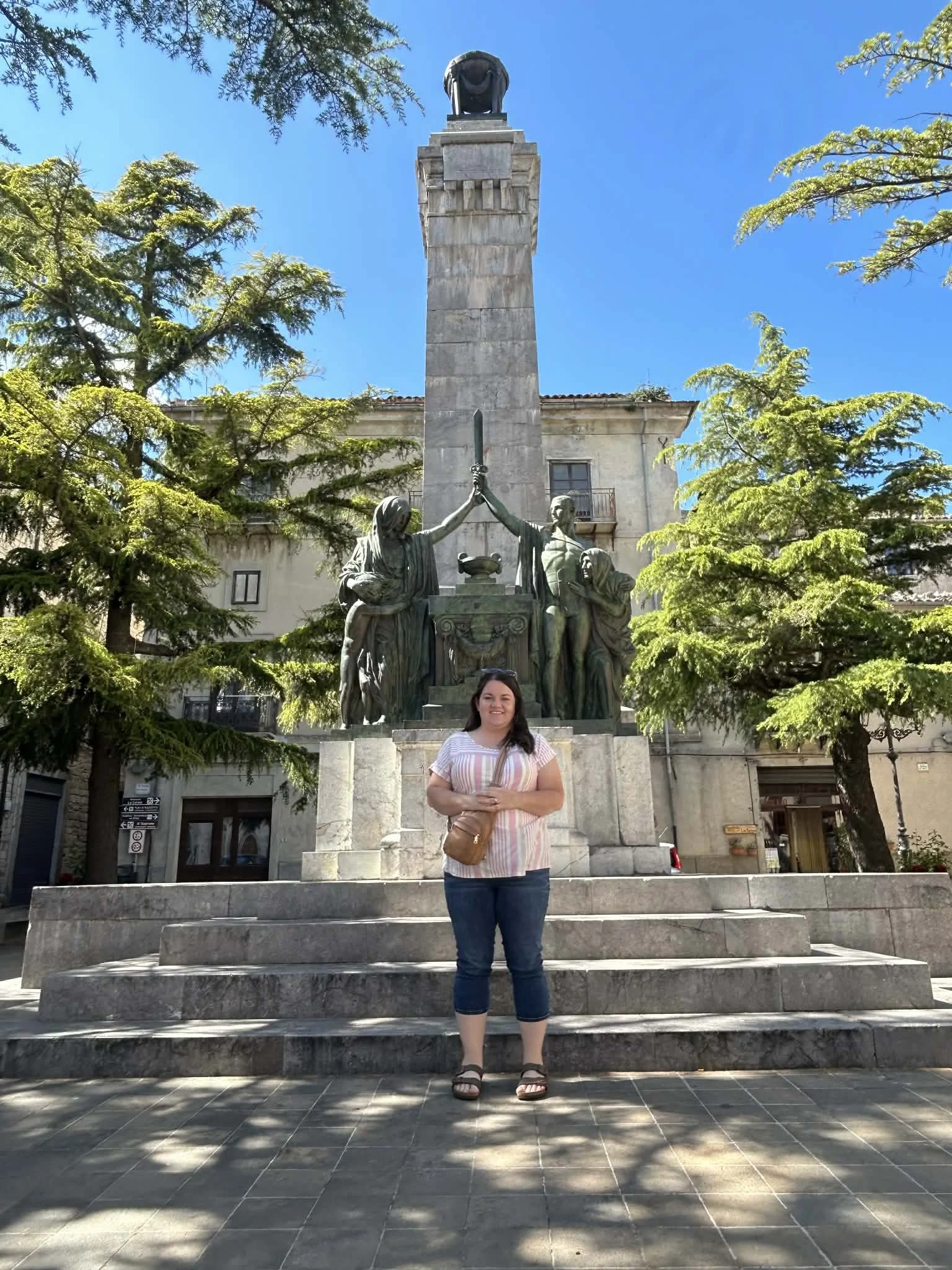 A woman standing in front of a statue with a tall obelisk and multiple figures holding a sword, surrounded by trees and a building, on a sunny day.