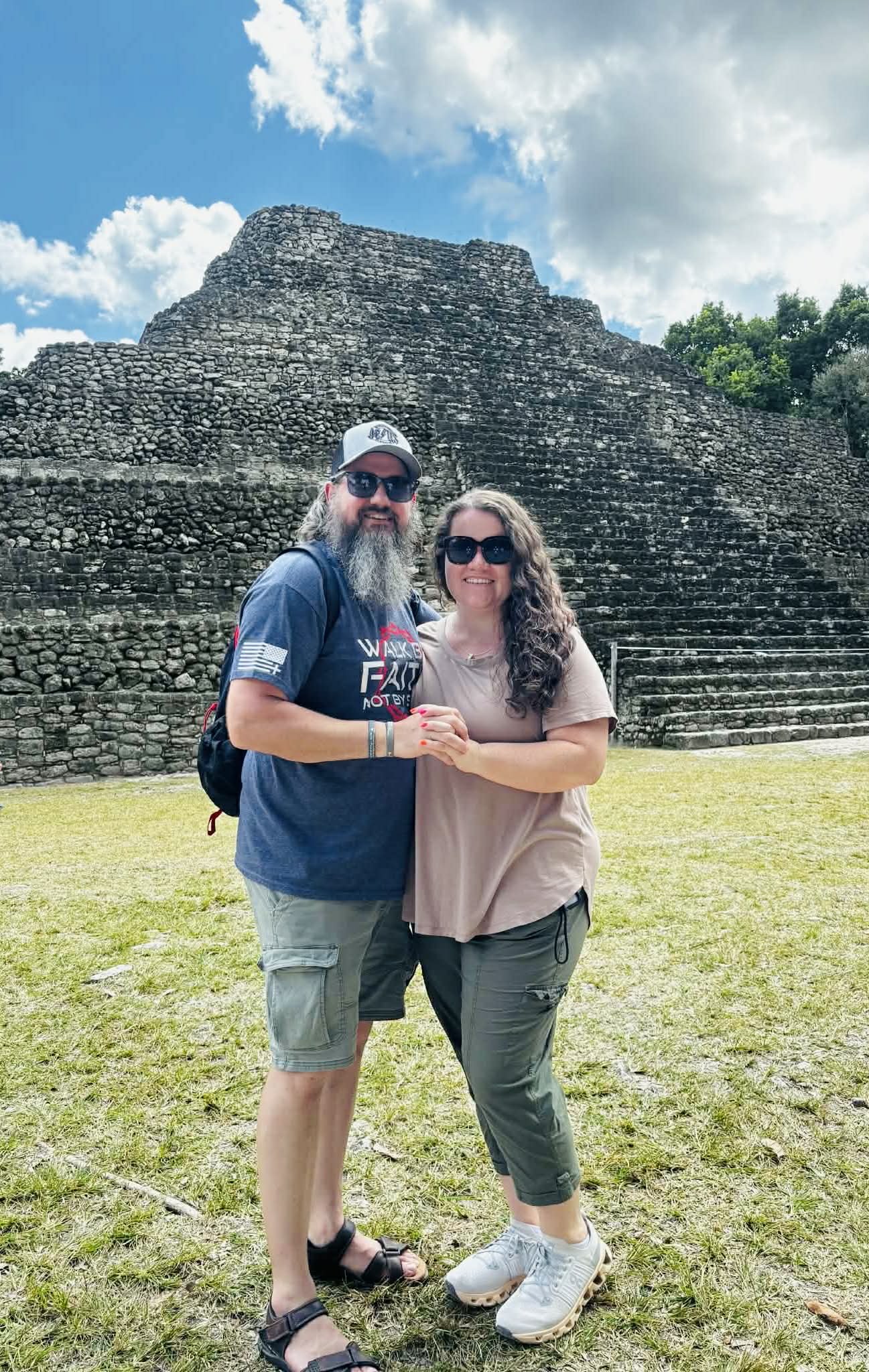 A couple standing in front of an ancient stepped stone pyramid at an archaeological site, smiling and holding hands.