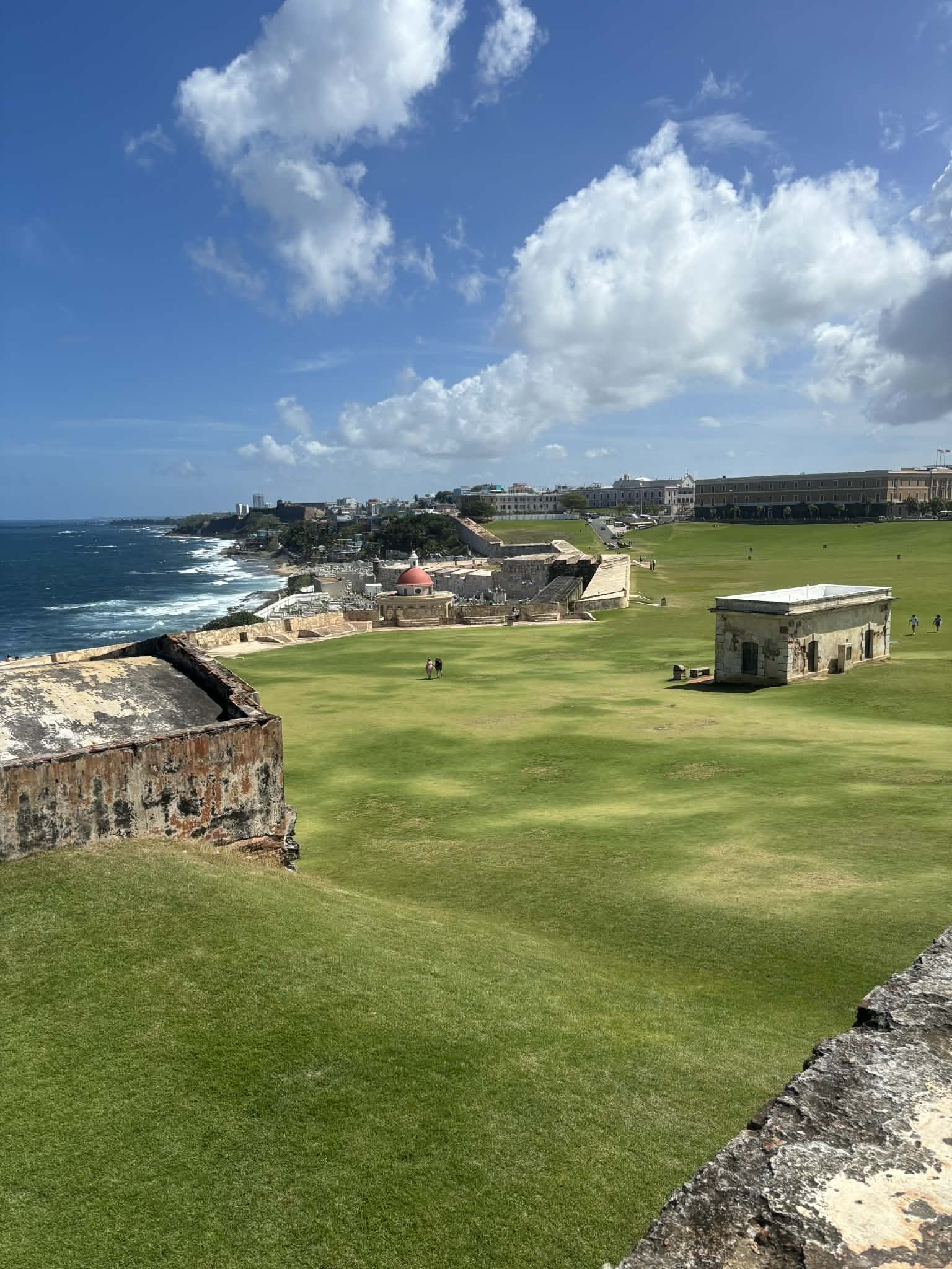 View of a historic fort with green grassy areas and old buildings, overlooking the coastline with waves and a partly cloudy sky.