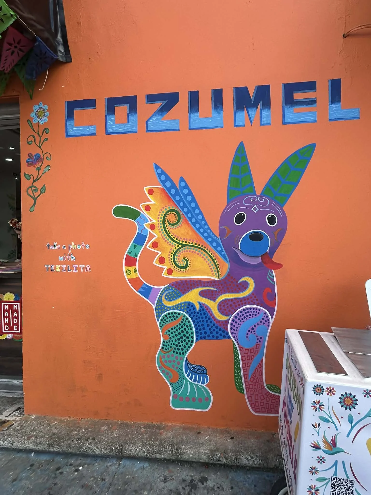 Colorful mural of a dog with dragonfly wings, decorated with vibrant patterns, on an orange wall at Cozumel.