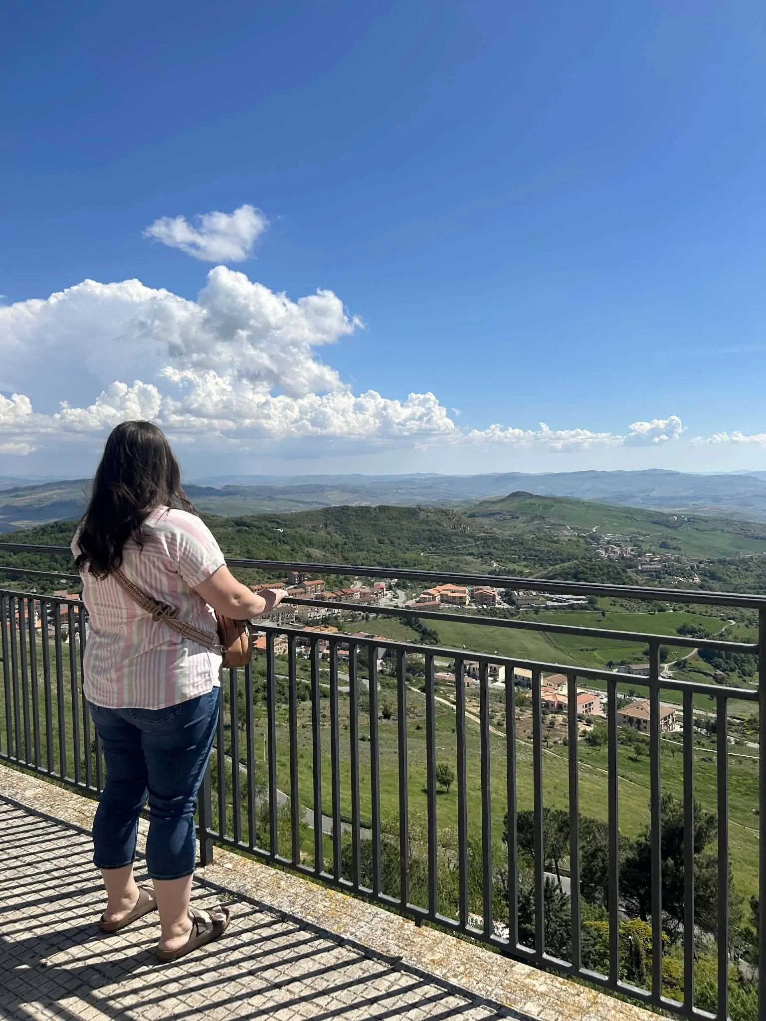 Woman standing on a balcony overlooking green hills and a small town under a blue sky with clouds.