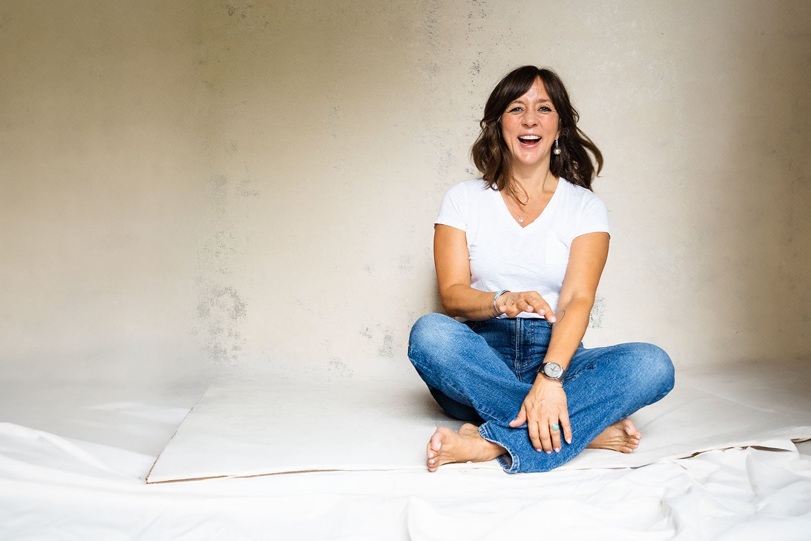 A woman sitting cross-legged on a white sheet against a plain beige wall, smiling and laughing, wearing a white T-shirt, blue jeans, and jewelry.