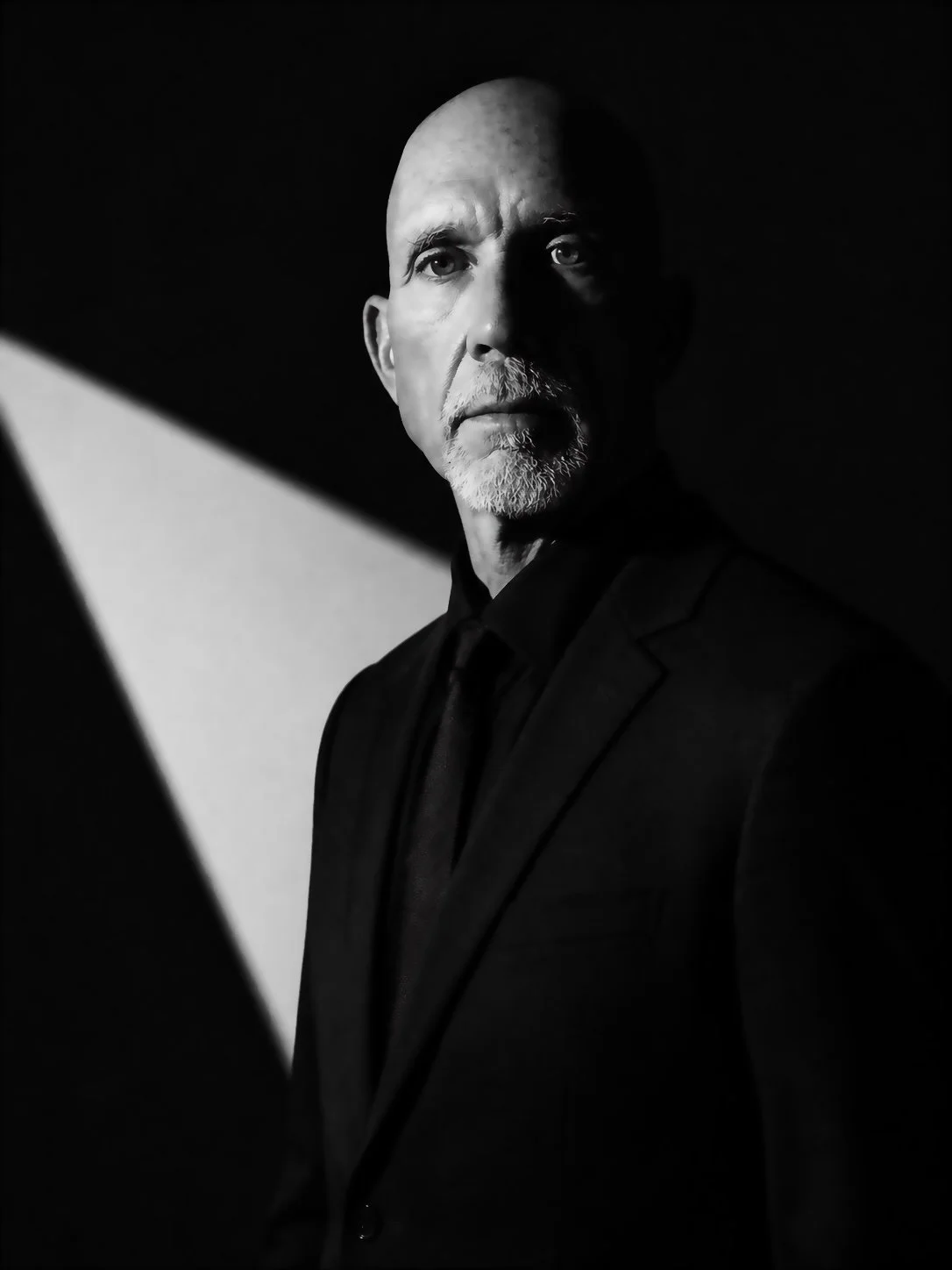 Black and white portrait of a serious-looking man with a beard and mustache, wearing a suit, with stark lighting creating high contrast and shadow effects.