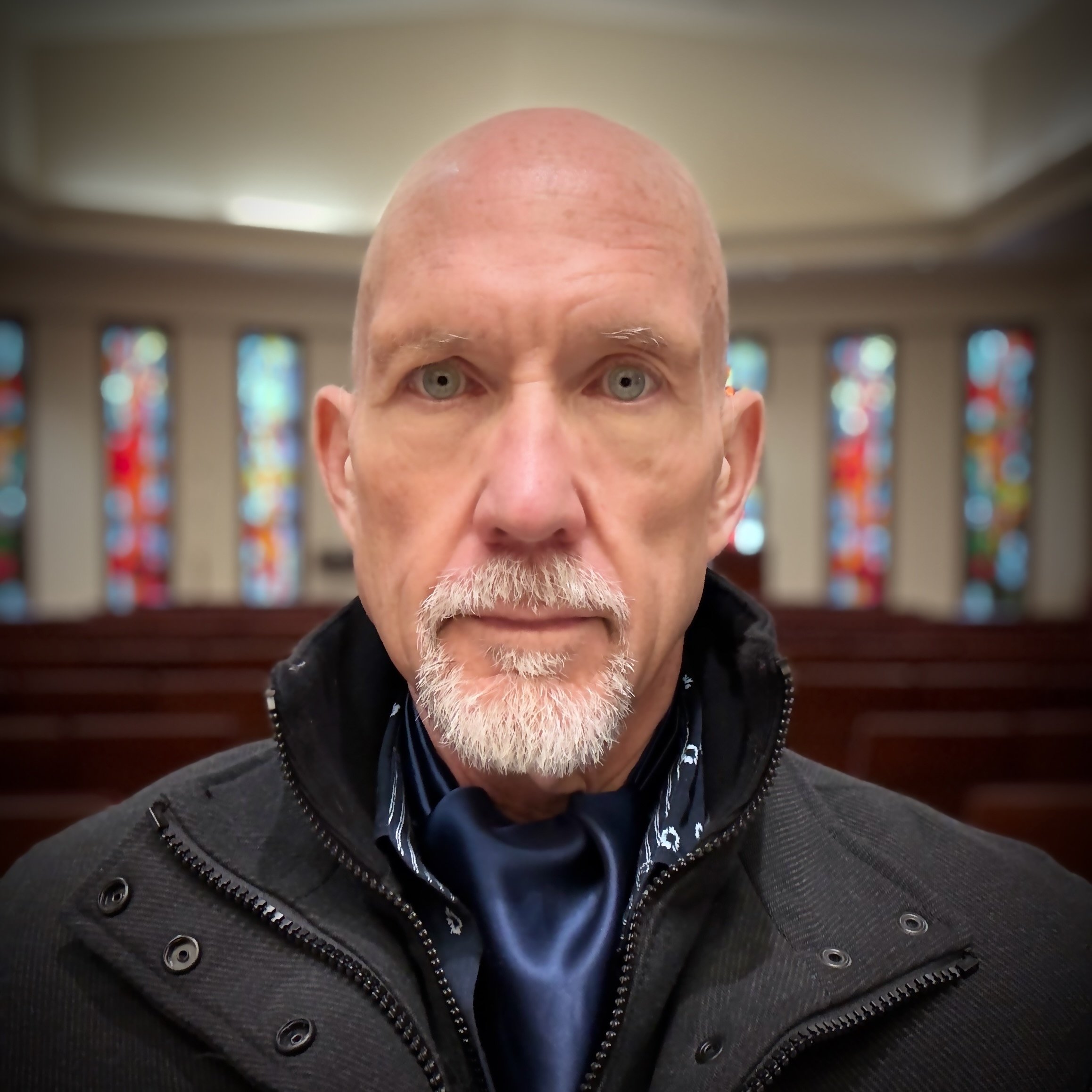 Close-up of bald man with gray beard, wearing a dark jacket with zipper, inside a church with stained glass windows in the background.