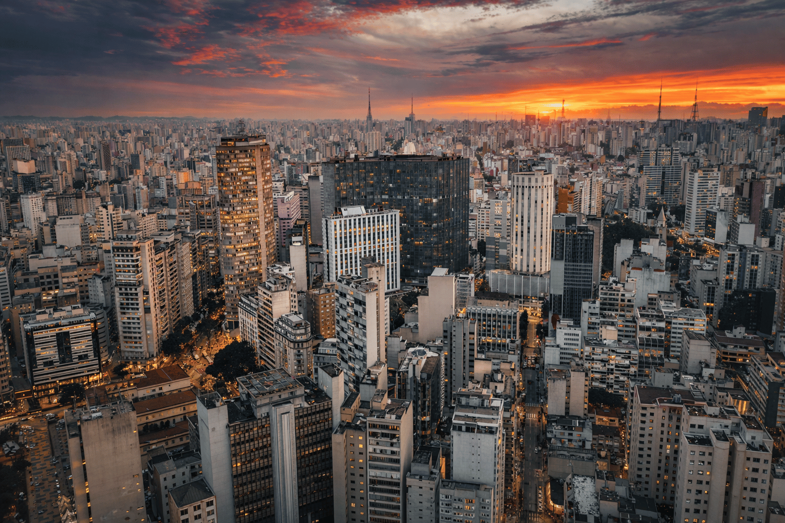 Vista aerial da cidade de São Paulo ao pôr do sol, com prédios altos e céu com nuvens coloridas
