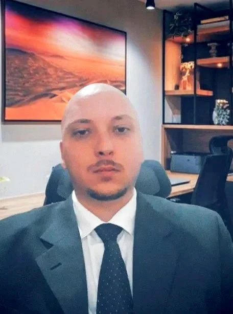 Homem de terno e gravata sentando em uma sala de escritório, com uma parede decorada com uma tela de arte abstrata e uma estante com livros e objetos ao fundo.