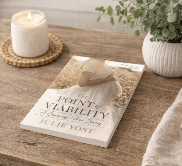 "The Point of Viability: A Coming Alive Story" book by Julie Yost on a wooden table next to a white candle and a potted green plant.