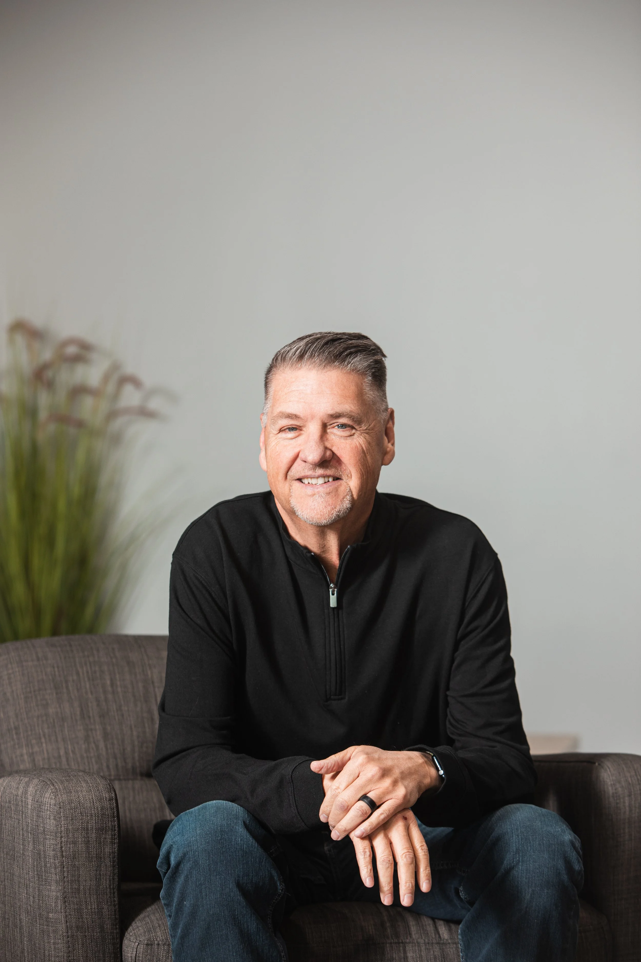 A smiling middle-aged man with short, gray hair and a beard, wearing a black zip-up jacket and blue jeans, sitting on a gray cushioned armchair with a dark surface in front. The background is a plain light gray wall with a few tall green plants on the left side.