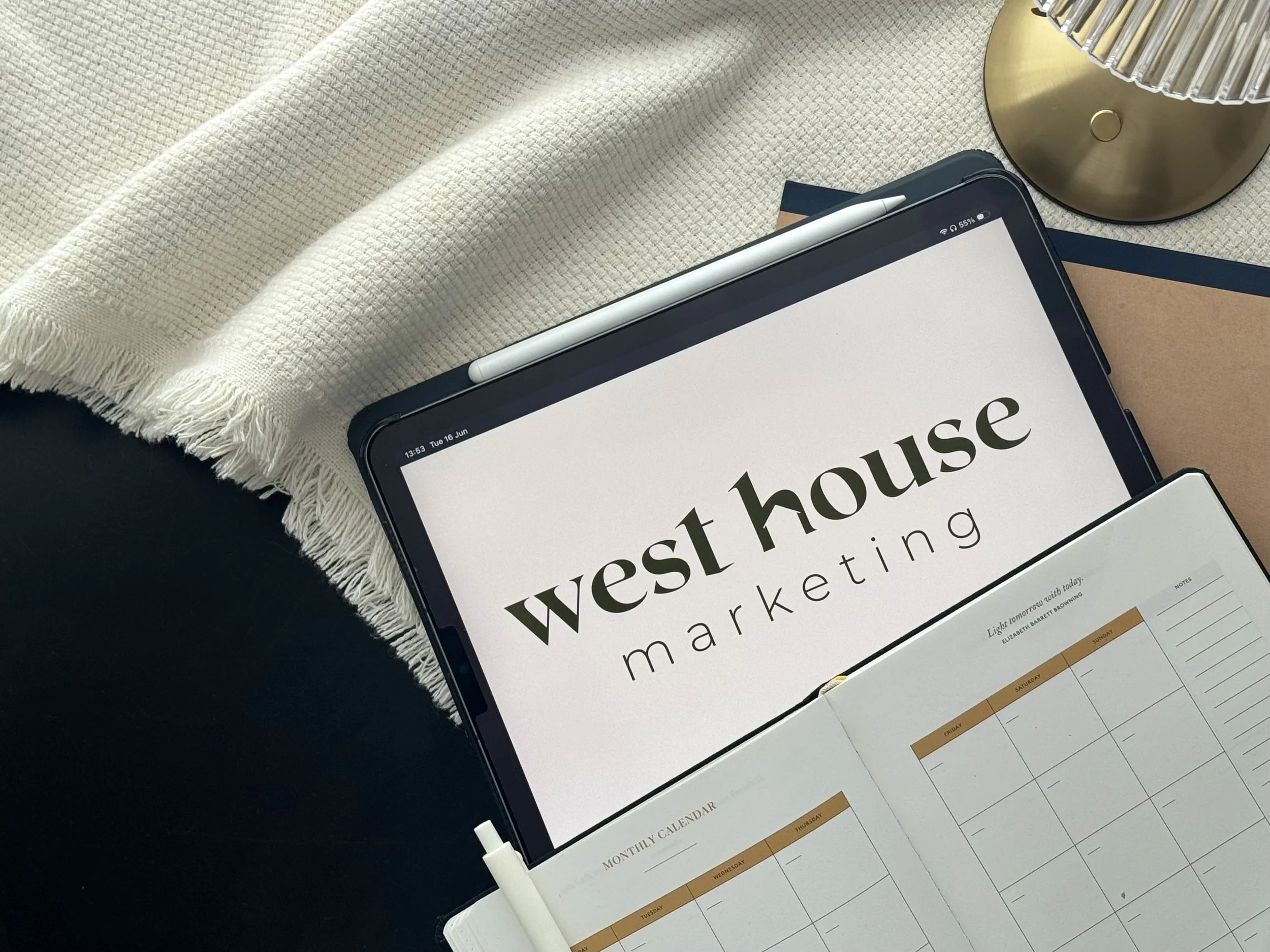 A tablet displaying the words 'post house' with part of a calendar below, a beige cloth with fringed edges, a gold lamp, and a stylus next to the tablet.