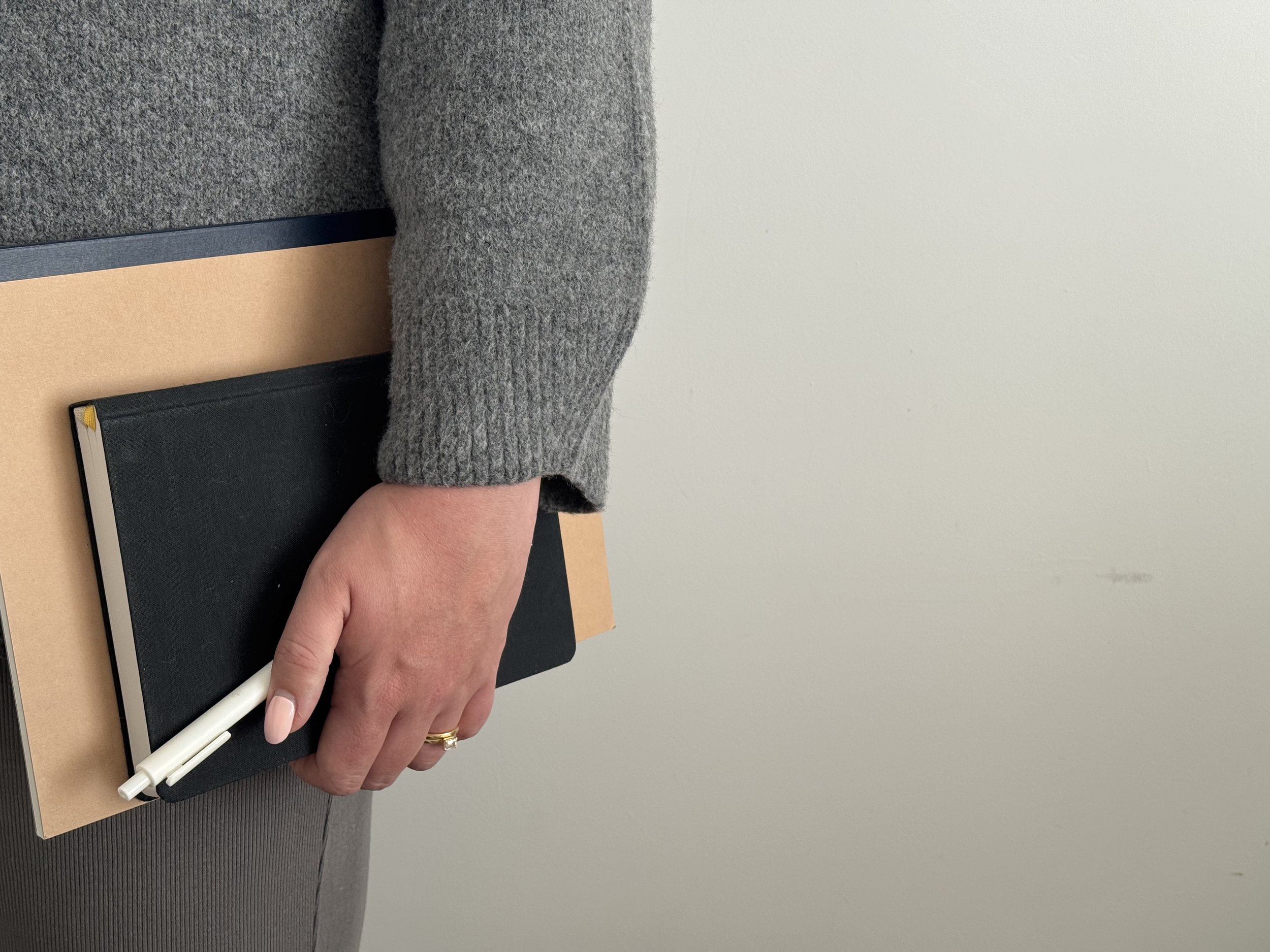 A person holding a notebook with a black cover and white pen, resting on a notebook with a tan cover, on a dark surface. The person is wearing a gray fuzzy sweater and has a gold ring on their left hand.