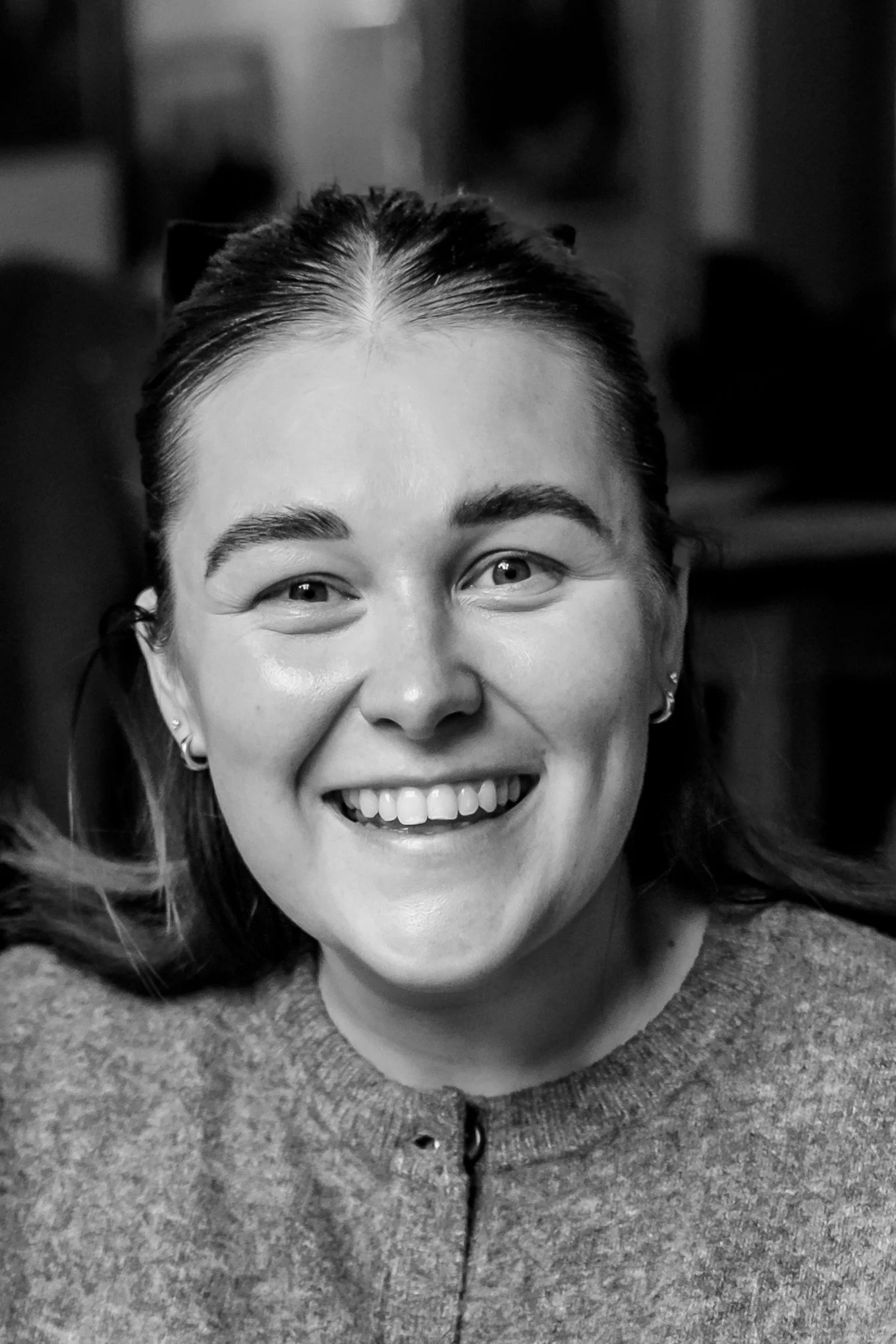 Close-up of a smiling woman with earrings, dark hair tied back, wearing a textured zip-up sweater, in black and white.