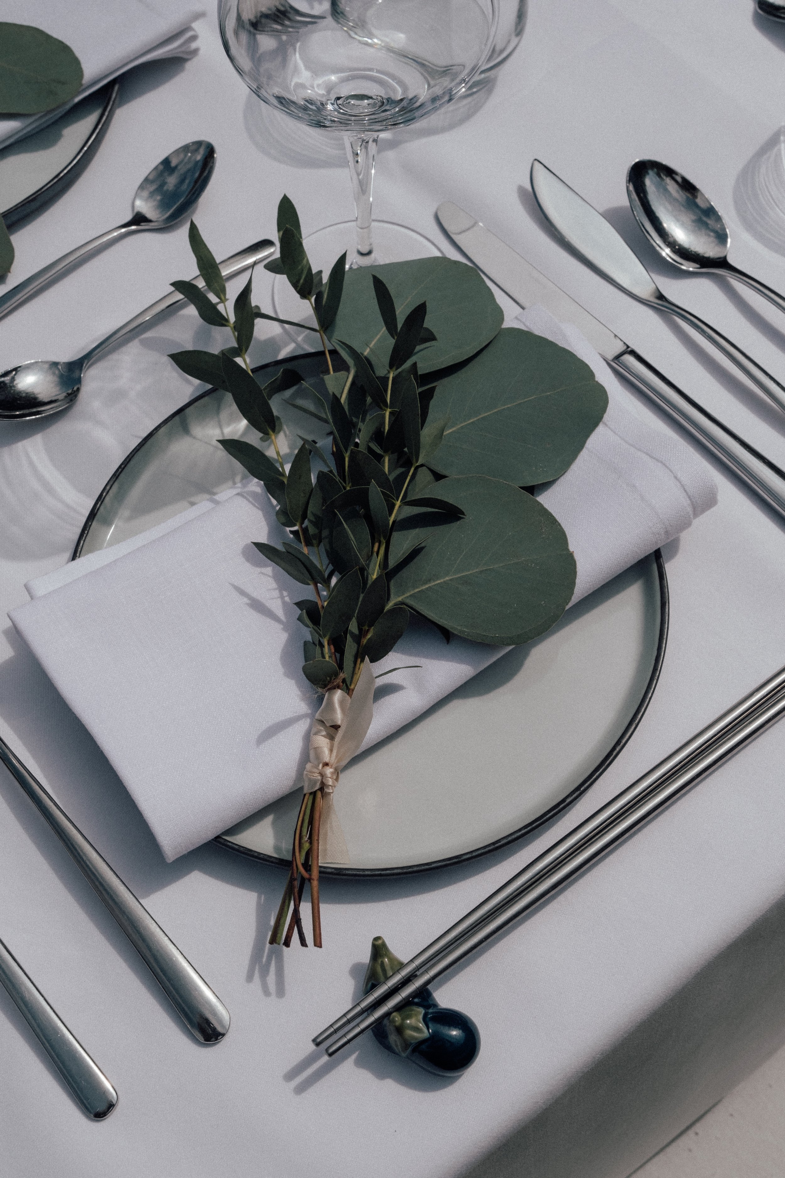 A table setting with silver utensils, a wine glass, a white napkin, and a small branch of greenery on a white tablecloth.