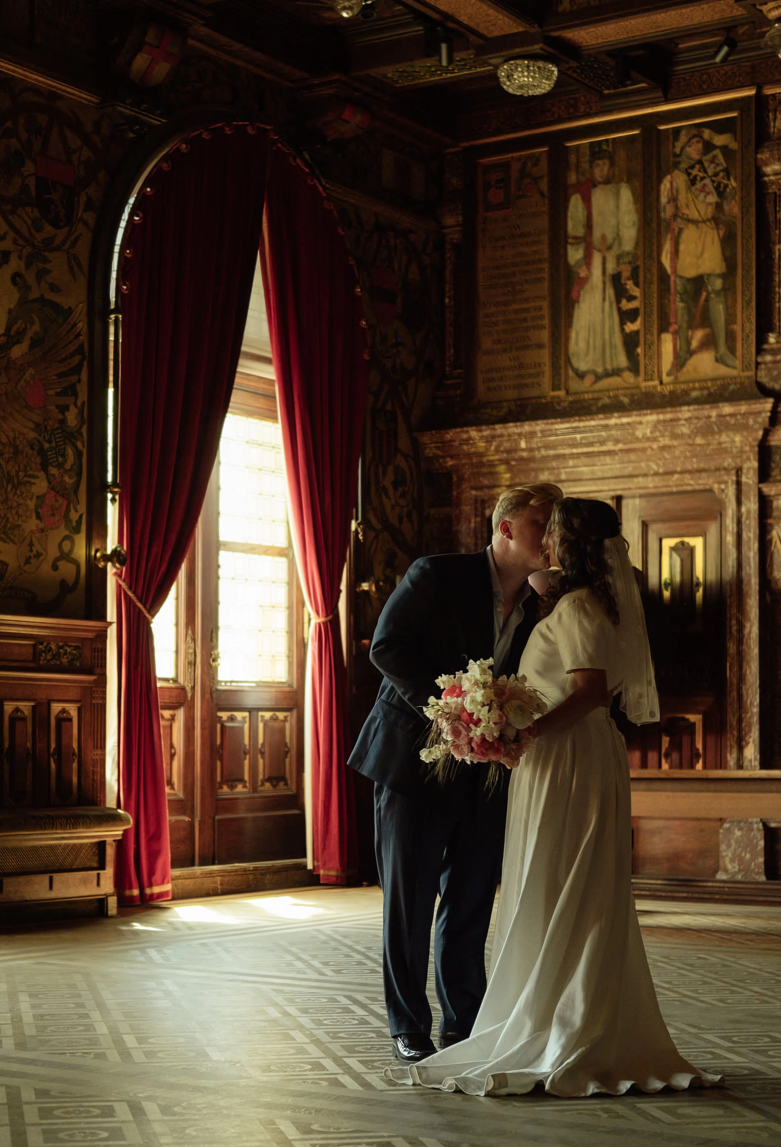 A bride and groom share a kiss during their wedding ceremony in a richly decorated wood-paneled room with large windows, red curtains, and historical portraits on the walls.