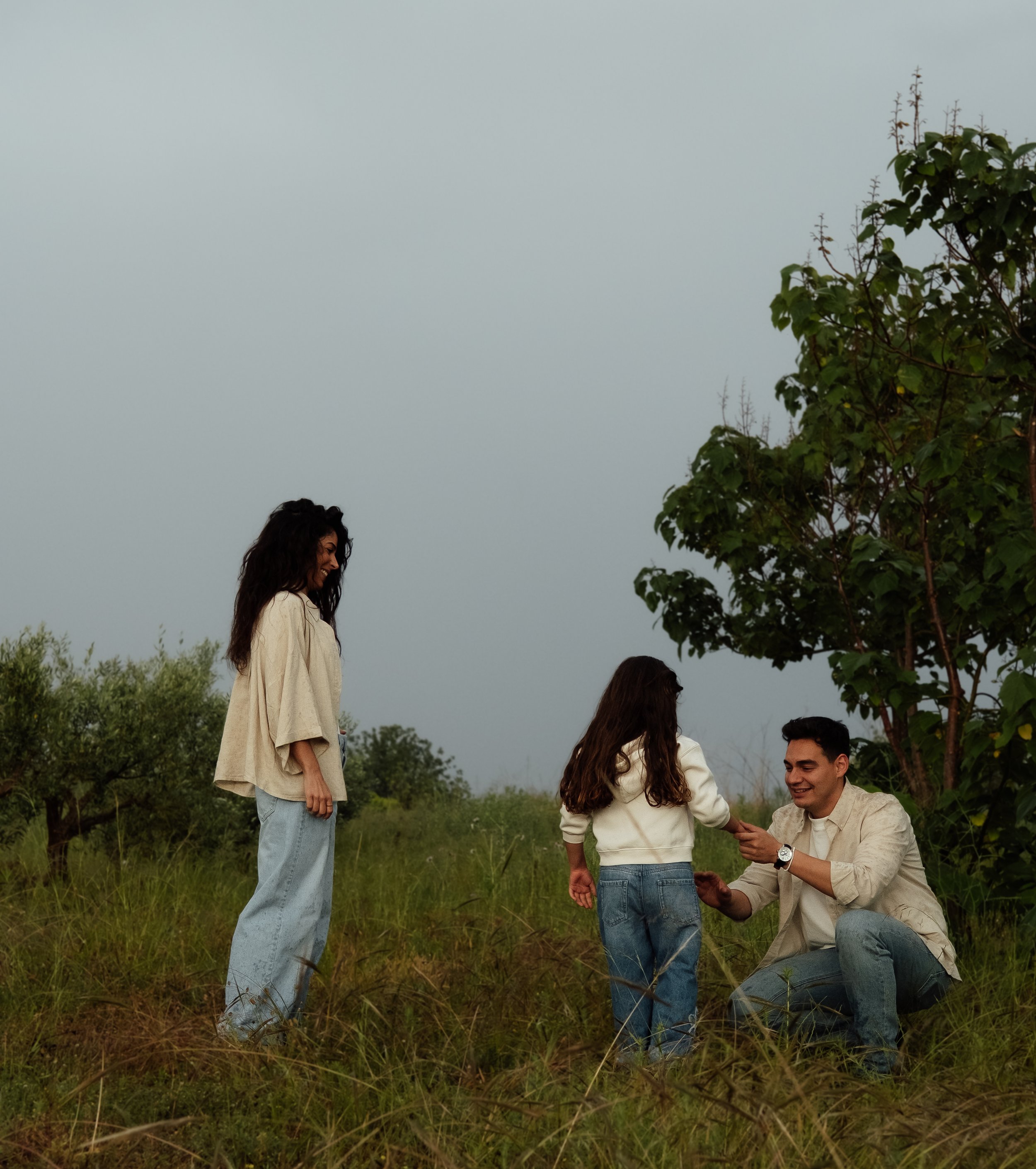 A group of four young adults with light to medium skin tones outdoors, with a cloudy sky and greenery around, sharing a moment of connection; three are standing, and one is kneeling, holding hands with another person.