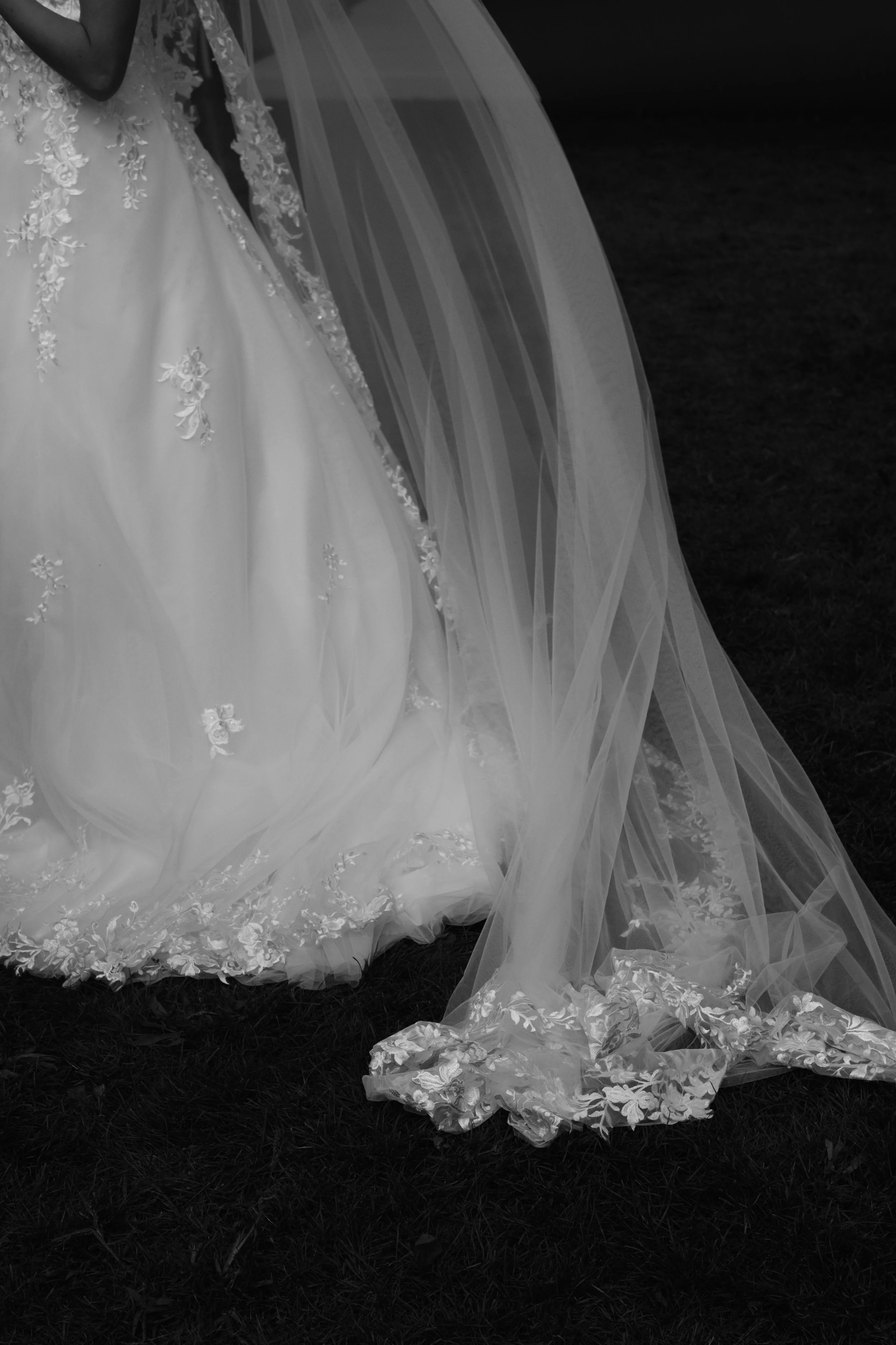Details of a white wedding dress with floral embroidery and a long, flowing veil lying on the grass.