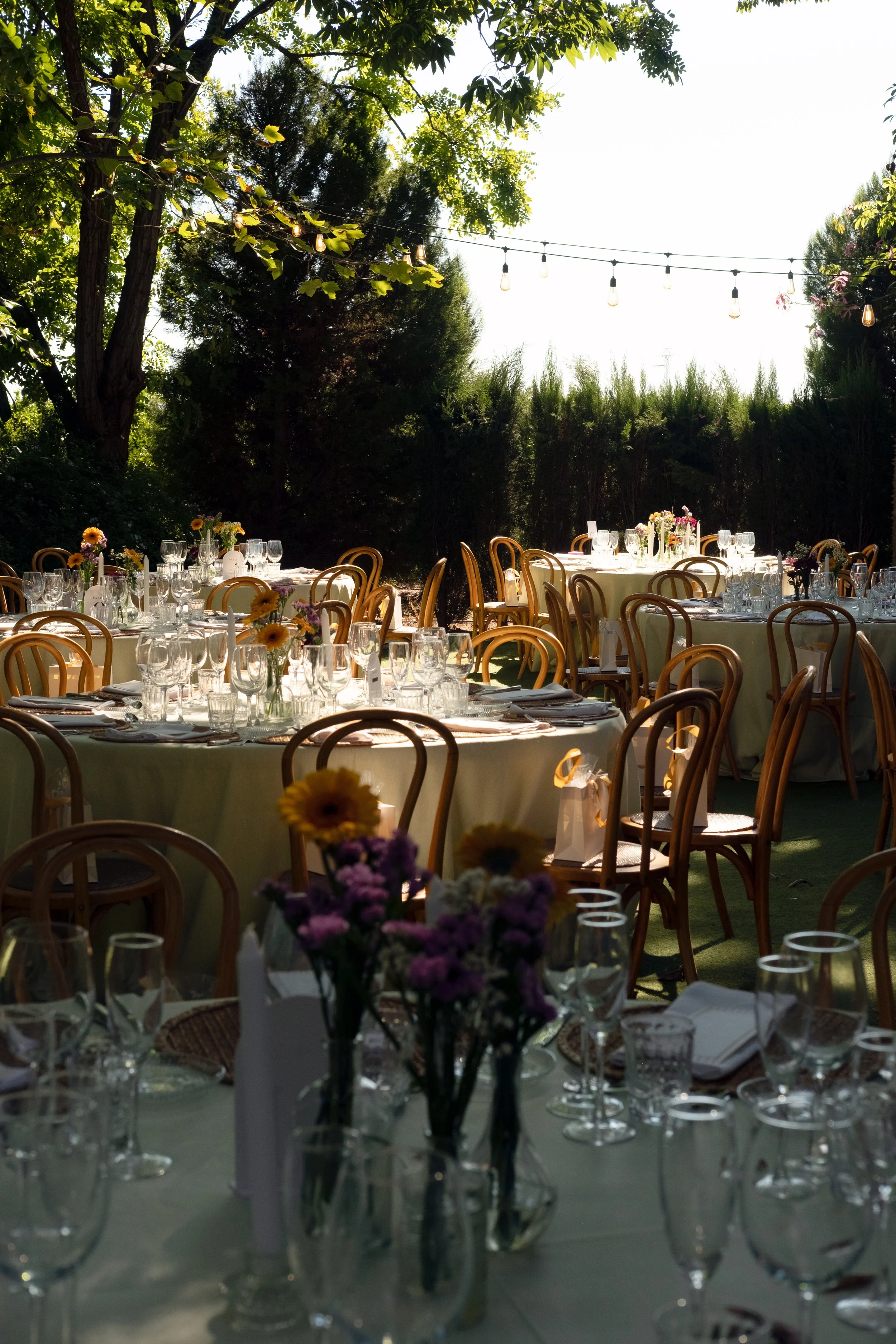 Outdoor dining setup with round tables covered in whitecloth, decorated with floral centerpieces, surrounded by wooden chairs, under string lights, with trees and bushes in the background.