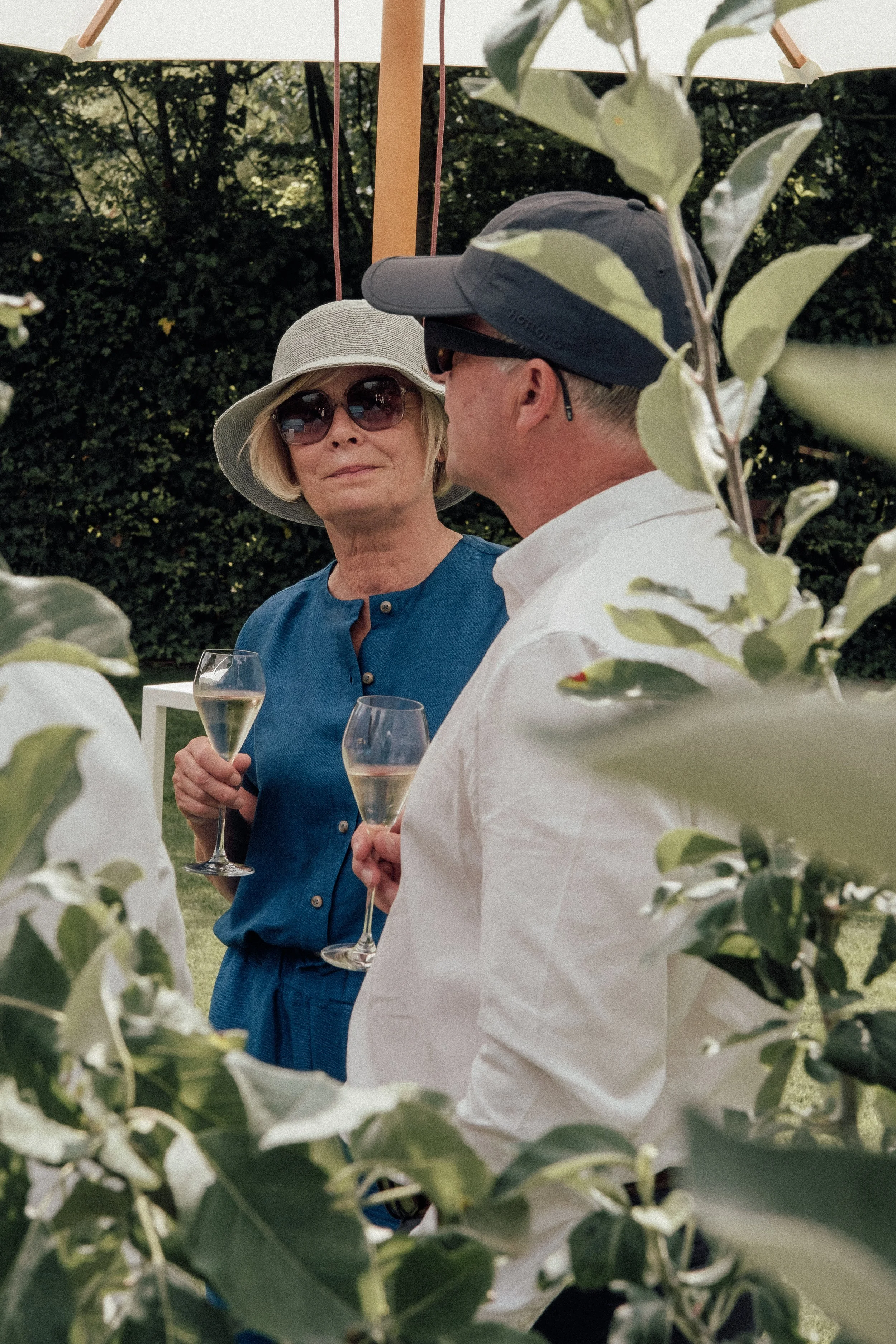 An elderly woman with blonde hair, wearing a wide-brimmed hat and sunglasses, holding a glass of white wine, talking to a man wearing a cap and sunglasses, at an outdoor gathering surrounded by greenery.